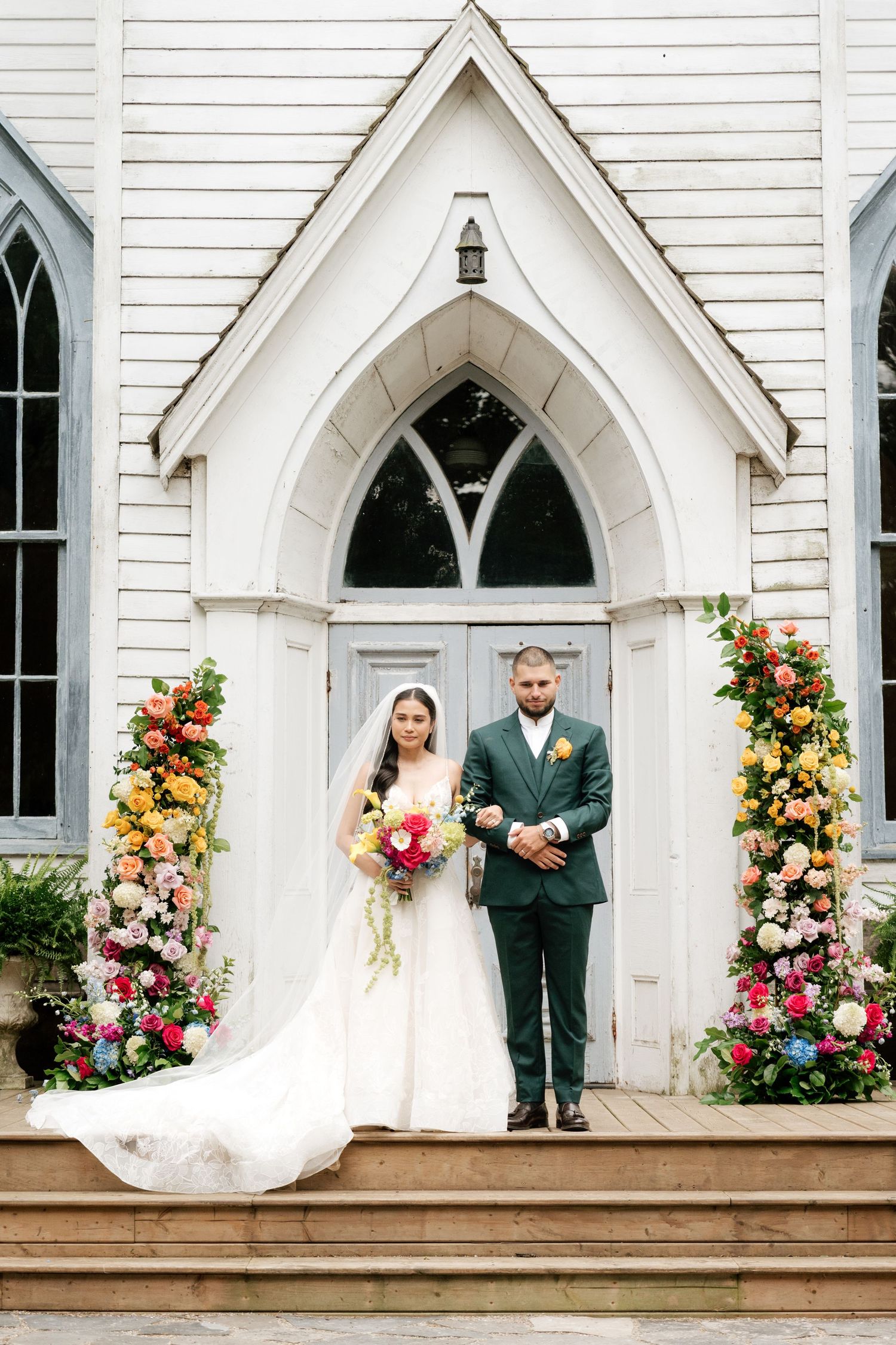 A wedding couple stands on the steps of a white Gothic church with arched doorways and floral arrangements.
