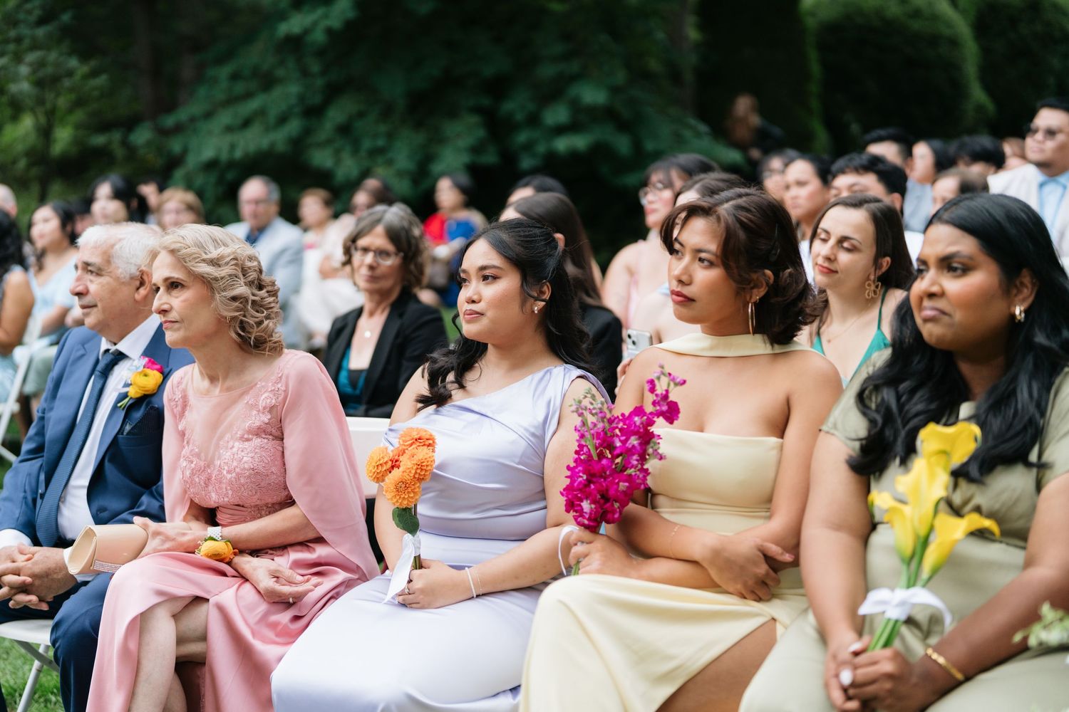Wedding guests seated outdoors react with joyful expressions during an emotional moment at the ceremony.