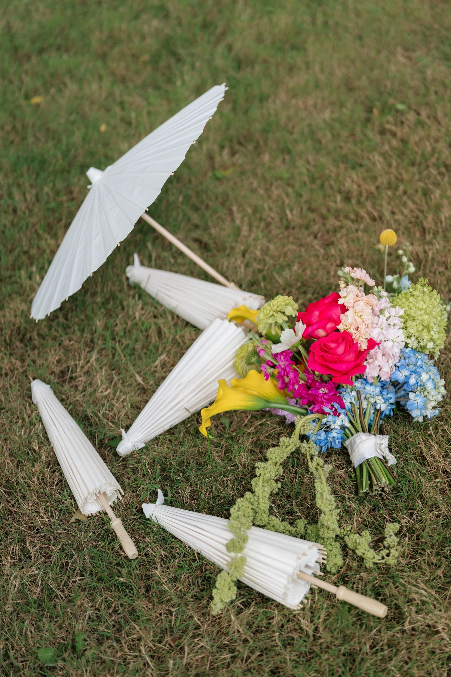 White paper parasols and a colorful bouquet with pink roses, yellow lilies, and blue hydrangeas laying on grass.