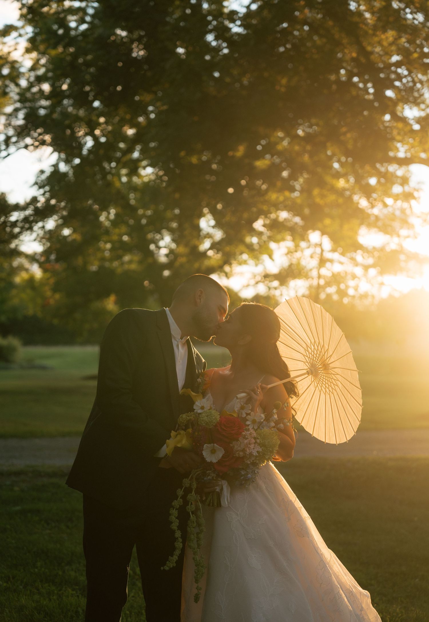 A bride and groom pose with decorative paper parasols during golden hour in an outdoor wedding setting.