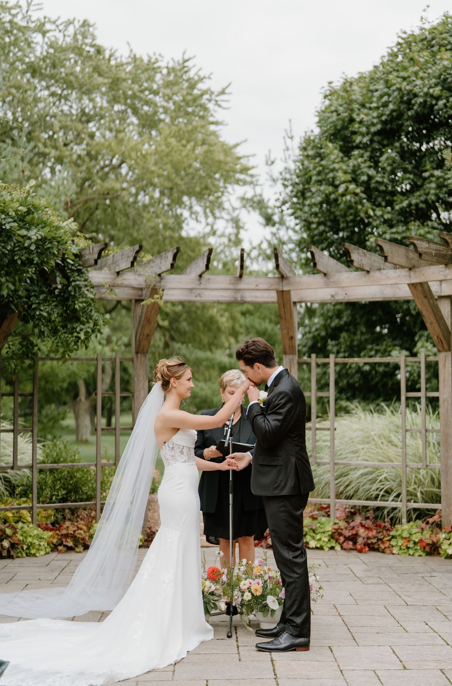 A couple shares an intimate moment during their wedding ceremony under a rustic wooden pergola surrounded by greenery.