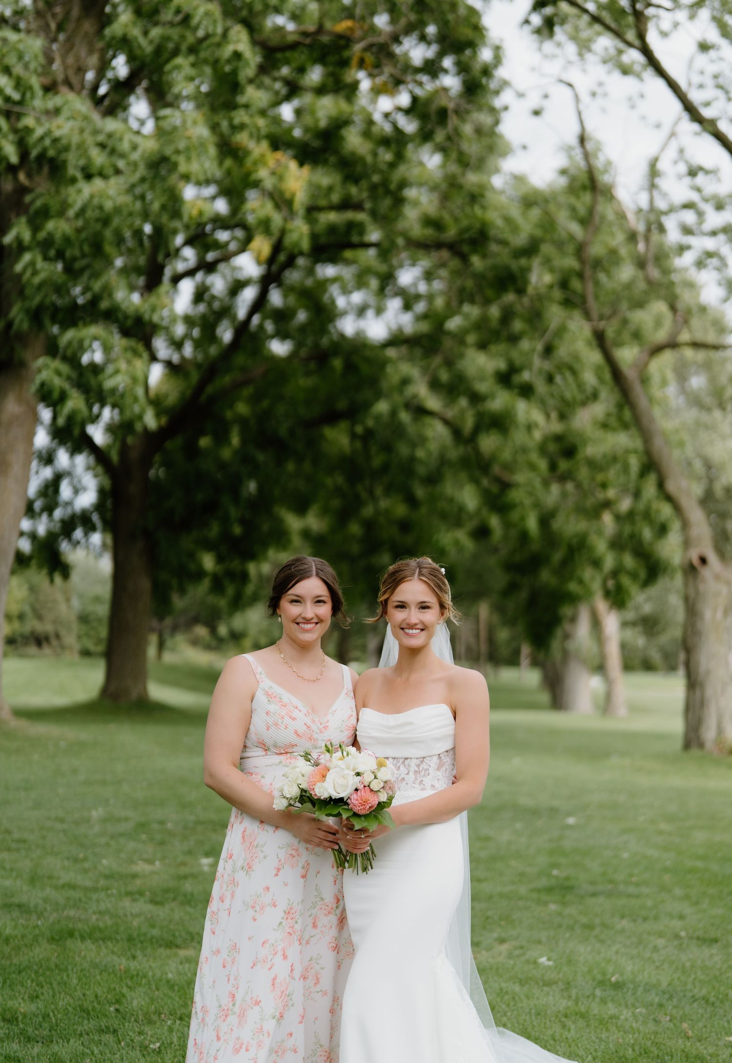 Wedding couple and bridesmaids in pastel dresses stand together on a lush green lawn surrounded by mature trees.