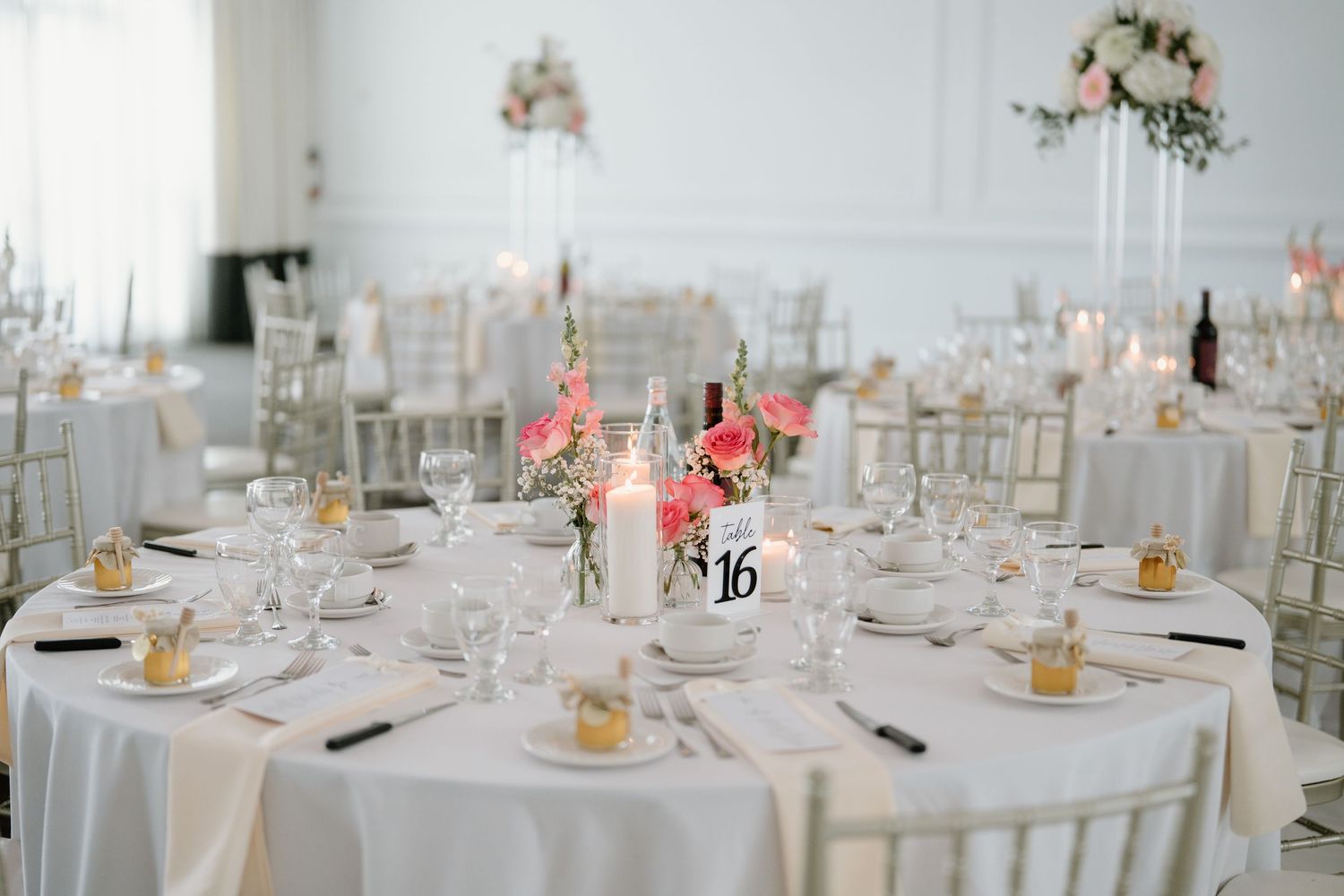 Elegant wedding reception tables with white linens, gold chairs, and tall coral floral centerpieces in a bright white room.
