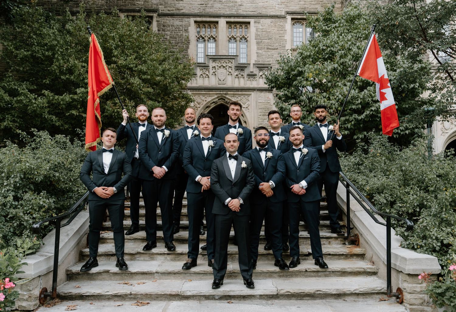 Groomsmen in black suits pose on stone steps for wedding photos with red flags in the background.