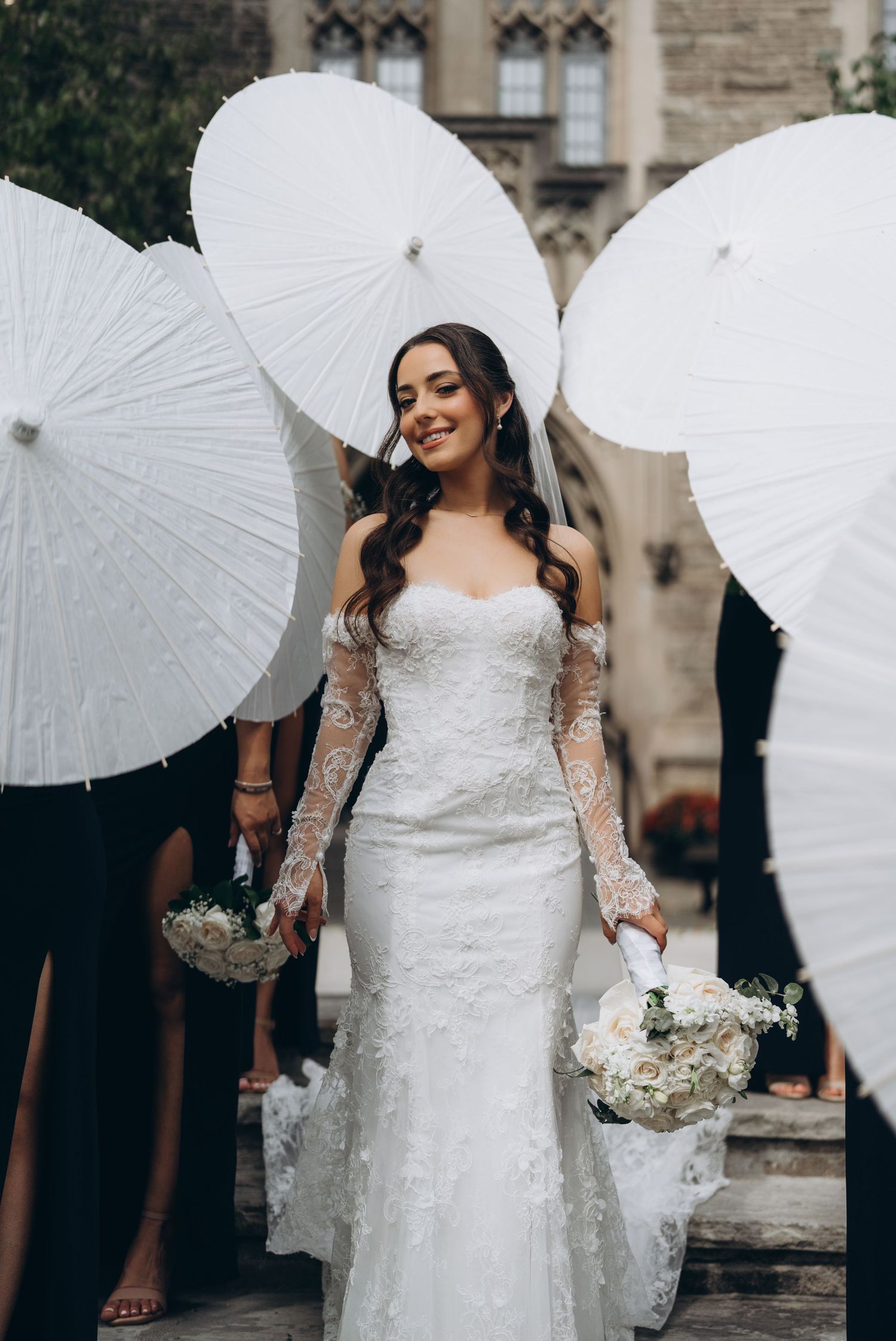 Bridal party holding white paper parasols poses together outside a stone building.