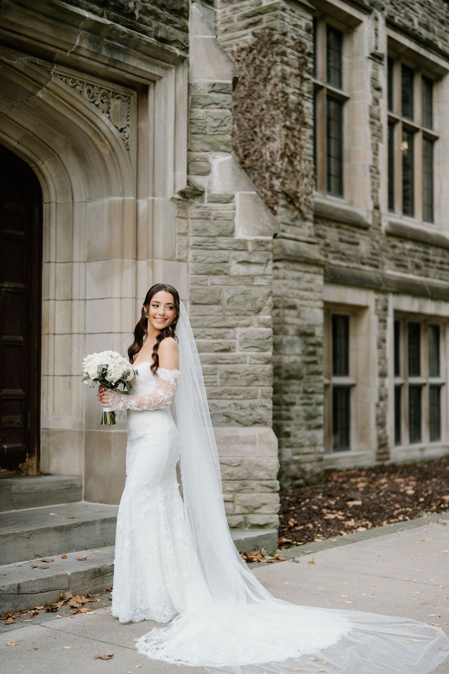 A bride in a white lace wedding dress poses elegantly against a stone building exterior.