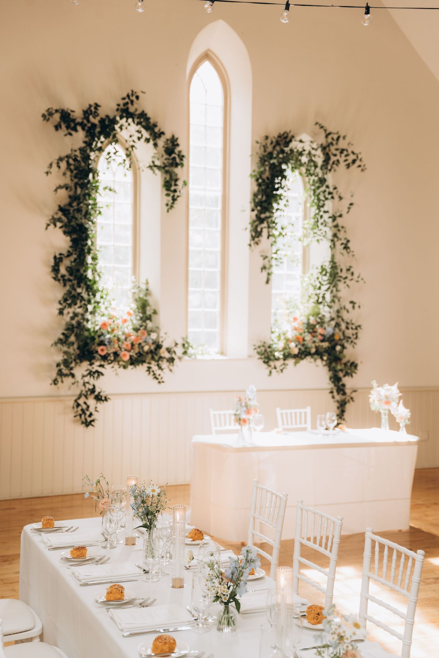 White minimalist wedding reception venue with arched windows, greenery decor, and white chairs at elegant dining tables.