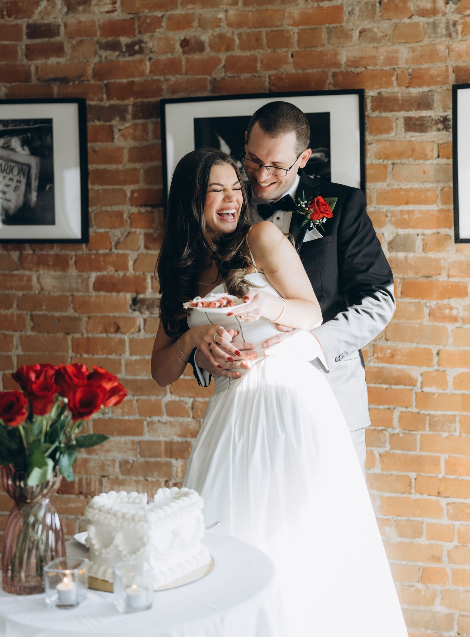 Joyful wedding couple shares a sweet moment cutting their cake together in front of a rustic brick backdrop.