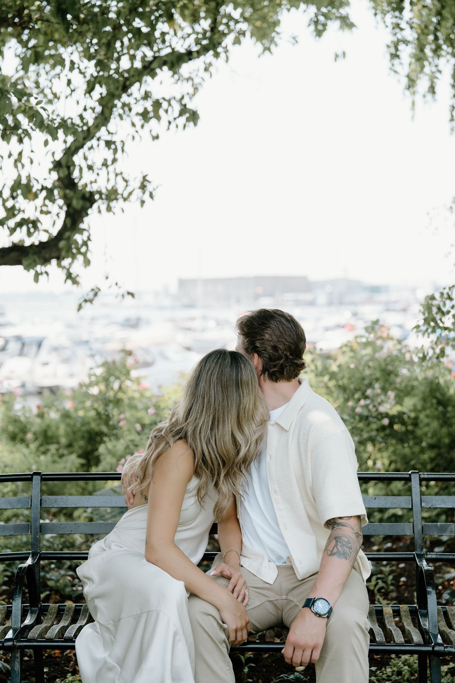 A couple sits intimately on a bench overlooking a scenic cityscape view framed by green tree branches.