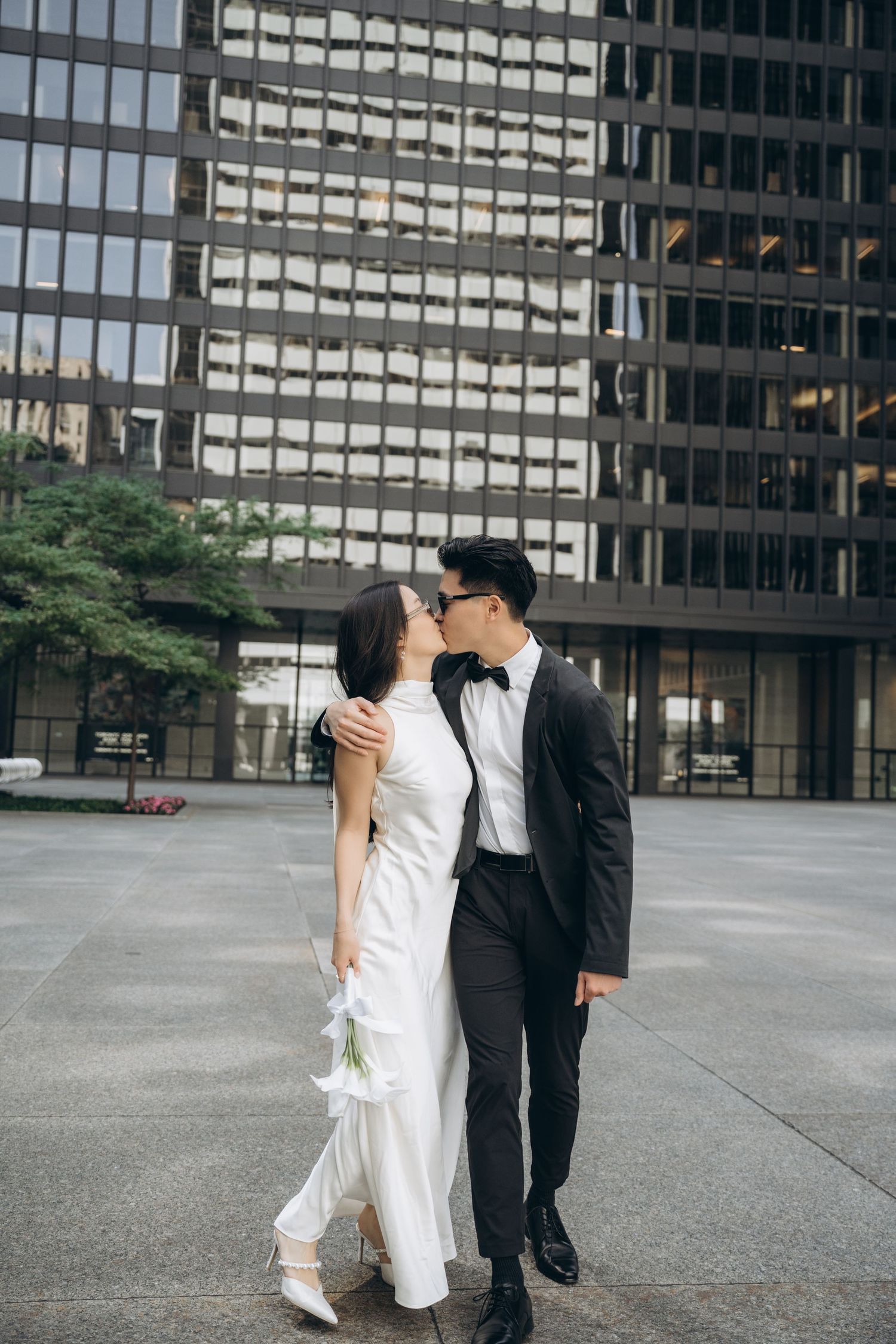 A couple in wedding attire share a romantic moment in front of modern glass office buildings in an urban plaza.