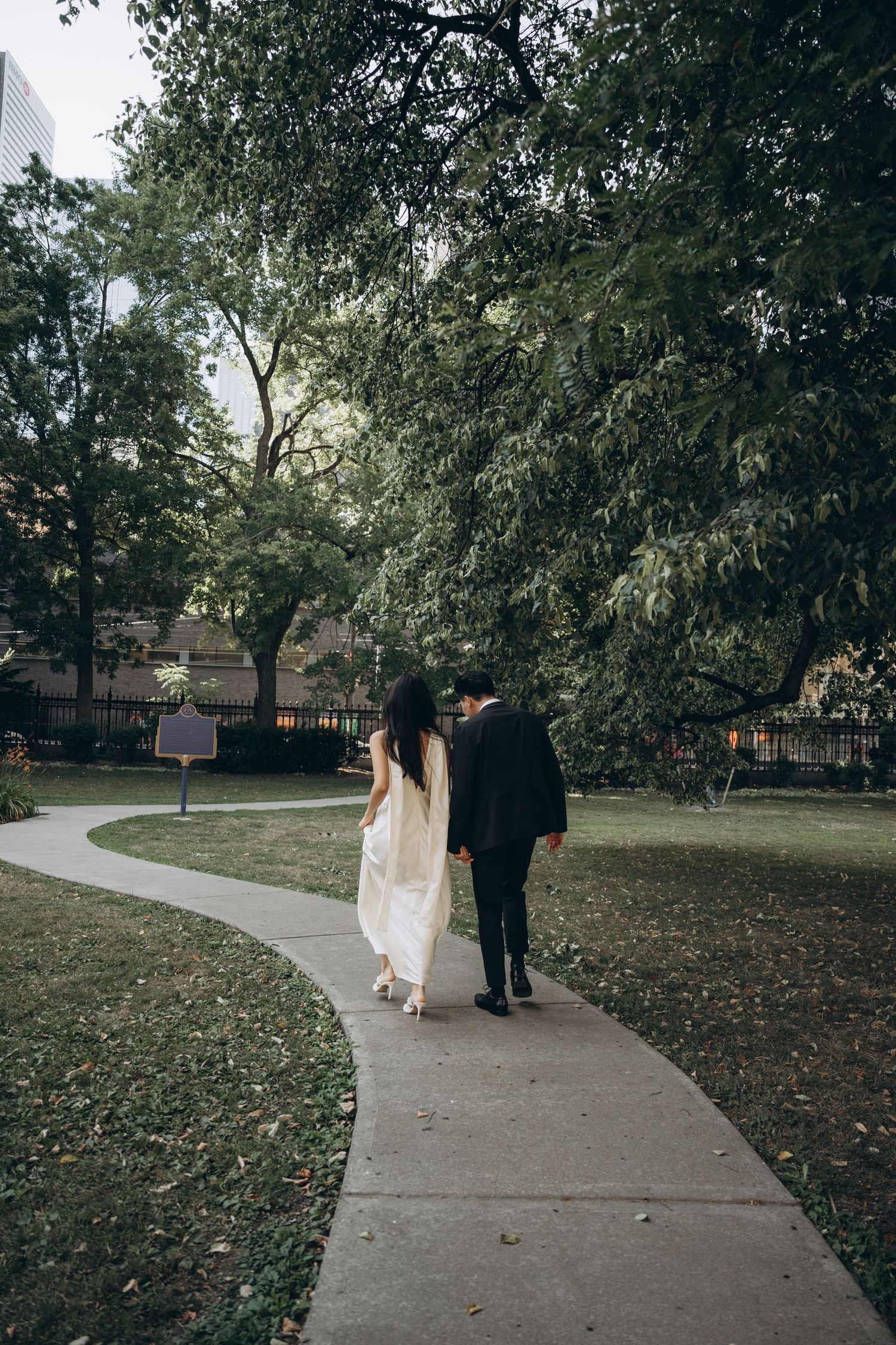 A couple walks together along a winding concrete path through a serene park with tall trees and scattered autumn leaves.