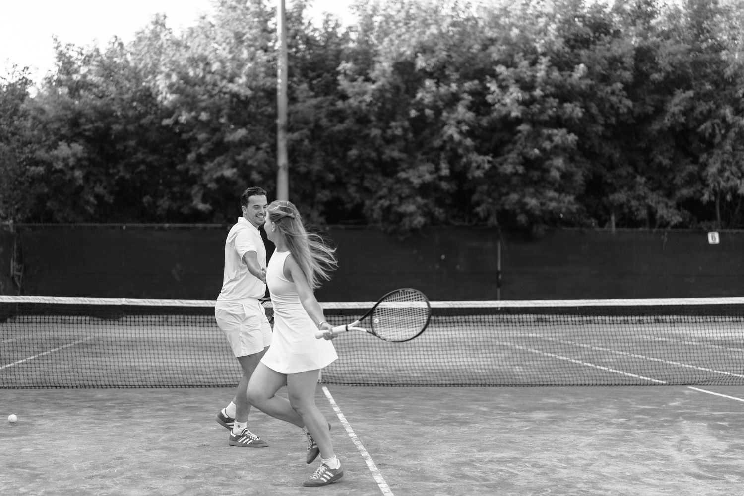 A black and white photo of a tennis player in action on a clay court with bushes in the background.