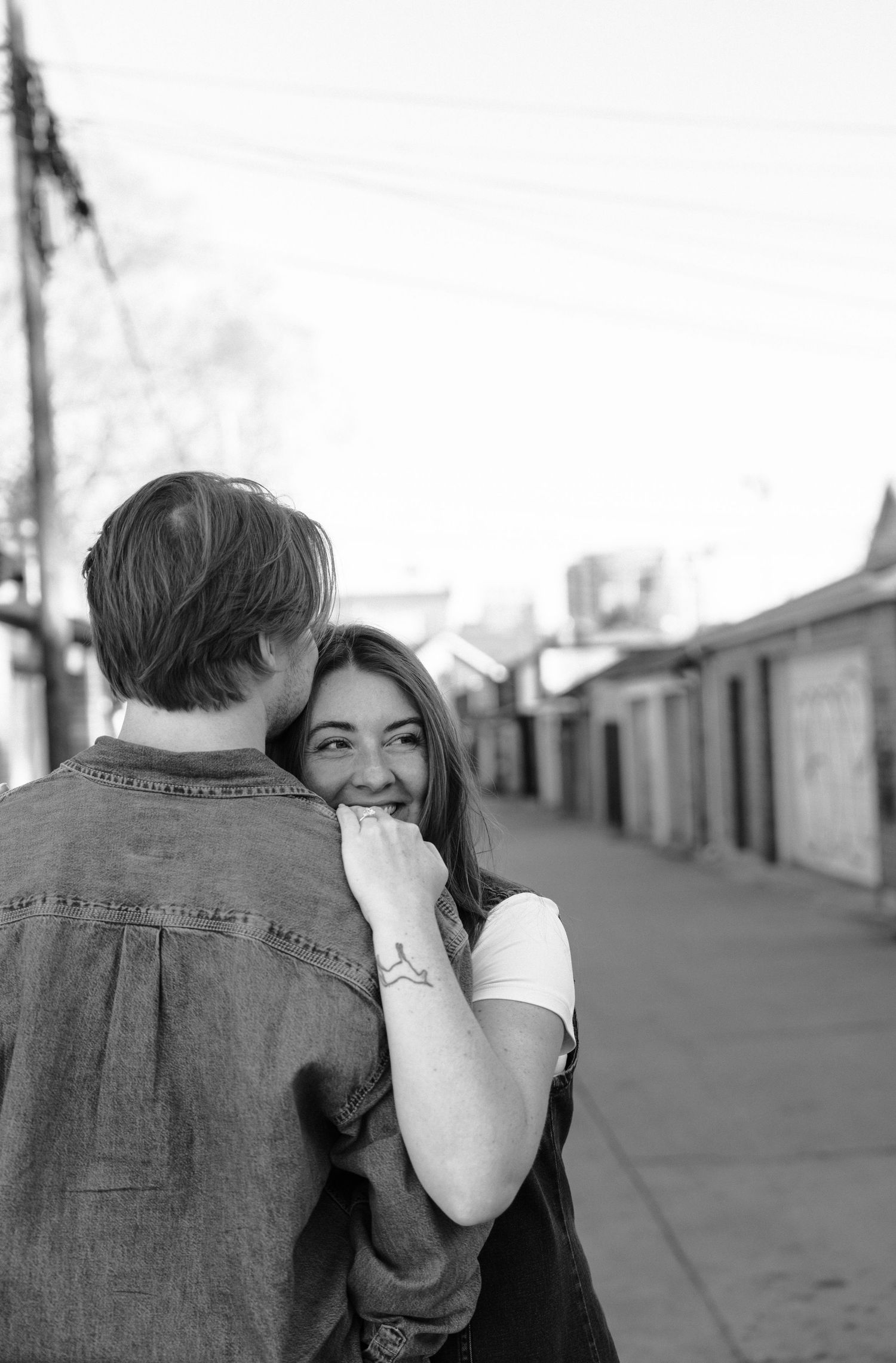 Black and white candid photograph of a romantic couple embracing on a quiet urban street with garages in the background.