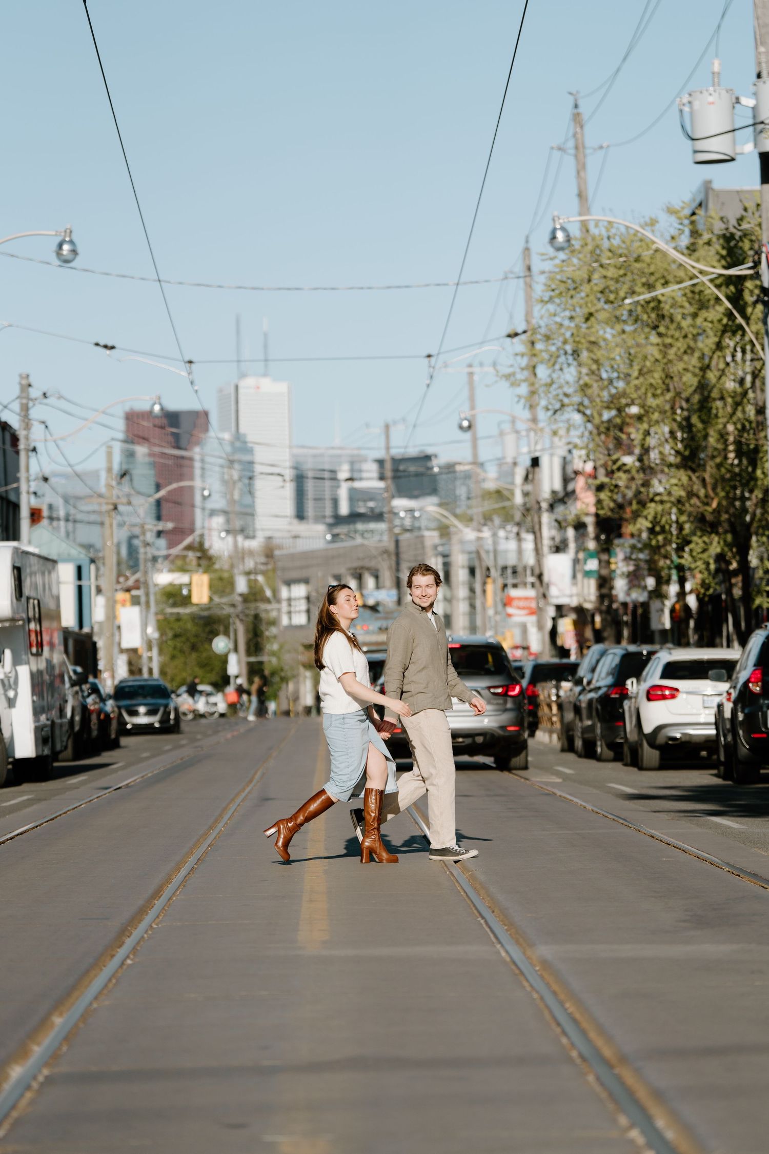 A couple stands on trolley tracks in an urban street with downtown buildings and utility lines visible in the background.
