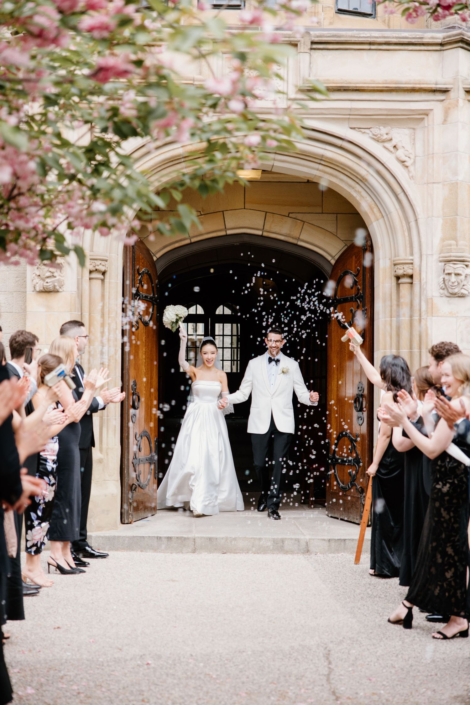 Wedding guests shower confetti on newlyweds exiting a stone church entrance lined with bridesmaids in black gowns.