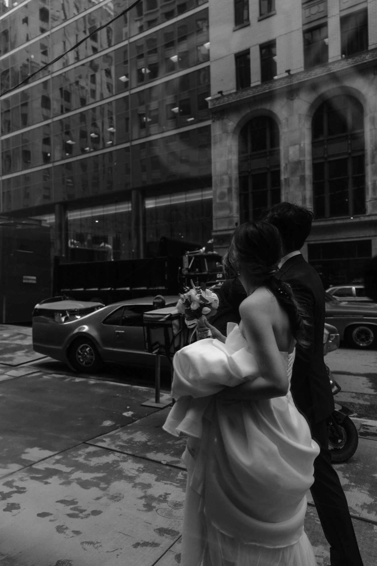 A couple in formal attire photographing on a city street with tall buildings and parked cars in black and white.
