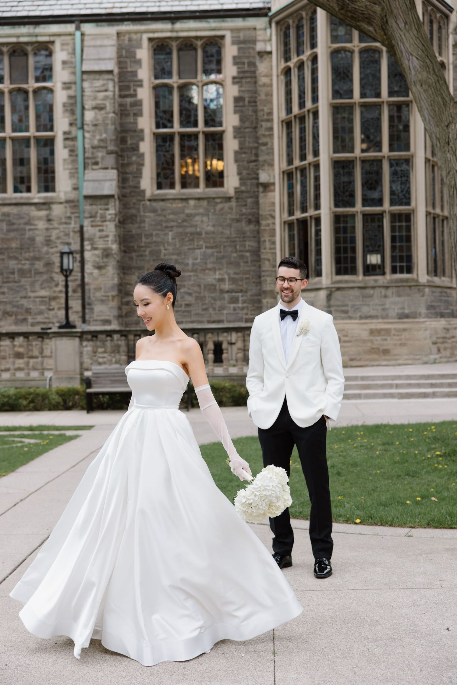 A bride in a classic white ballgown and groom in white tuxedo jacket walk along a stone building's pathway.