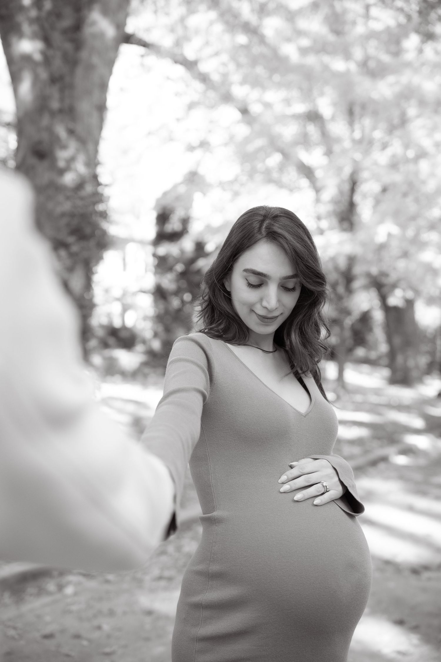 A series of black and white photos show a pregnant woman in a casual dress standing in a sunlit wooded area.