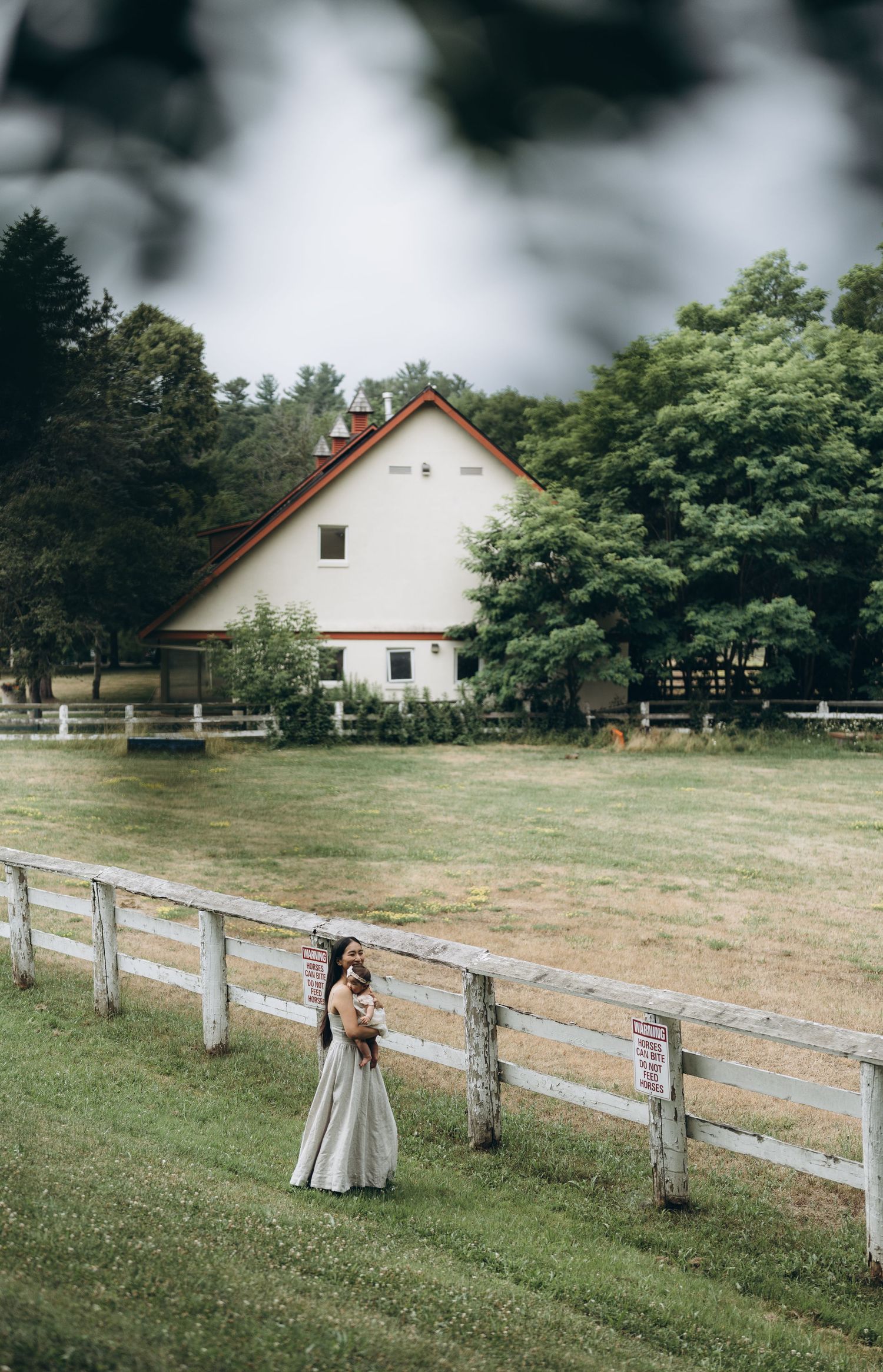 A white barn with red trim sits on a grassy property with wooden fence posts and lush trees in the background.