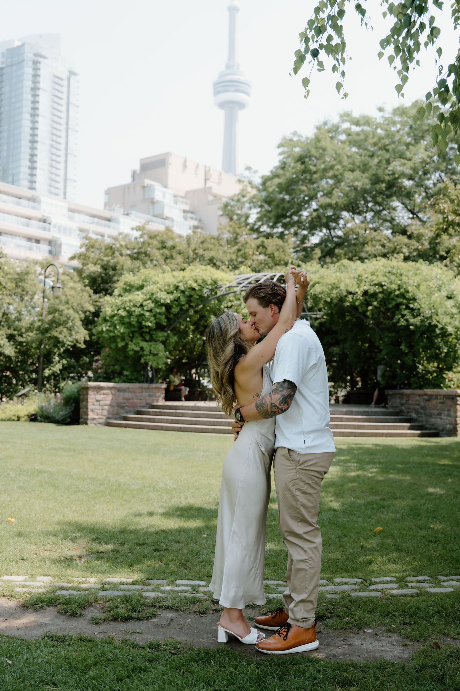 A romantic moment in a Toronto park with the CN Tower visible in the background on a sunny summer day.