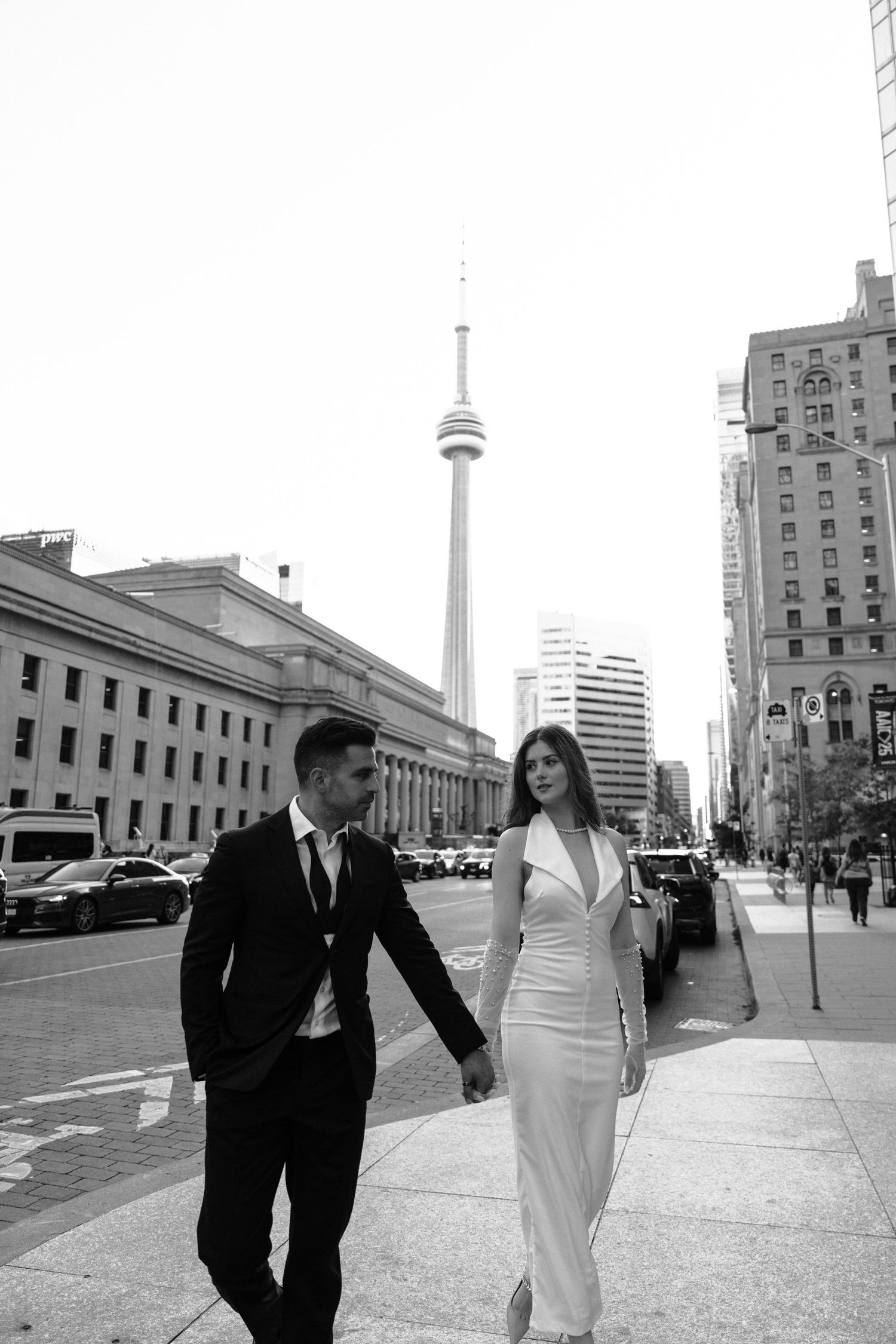 A couple walking on a Toronto sidewalk in formal attire with the CN Tower and downtown buildings visible in background.