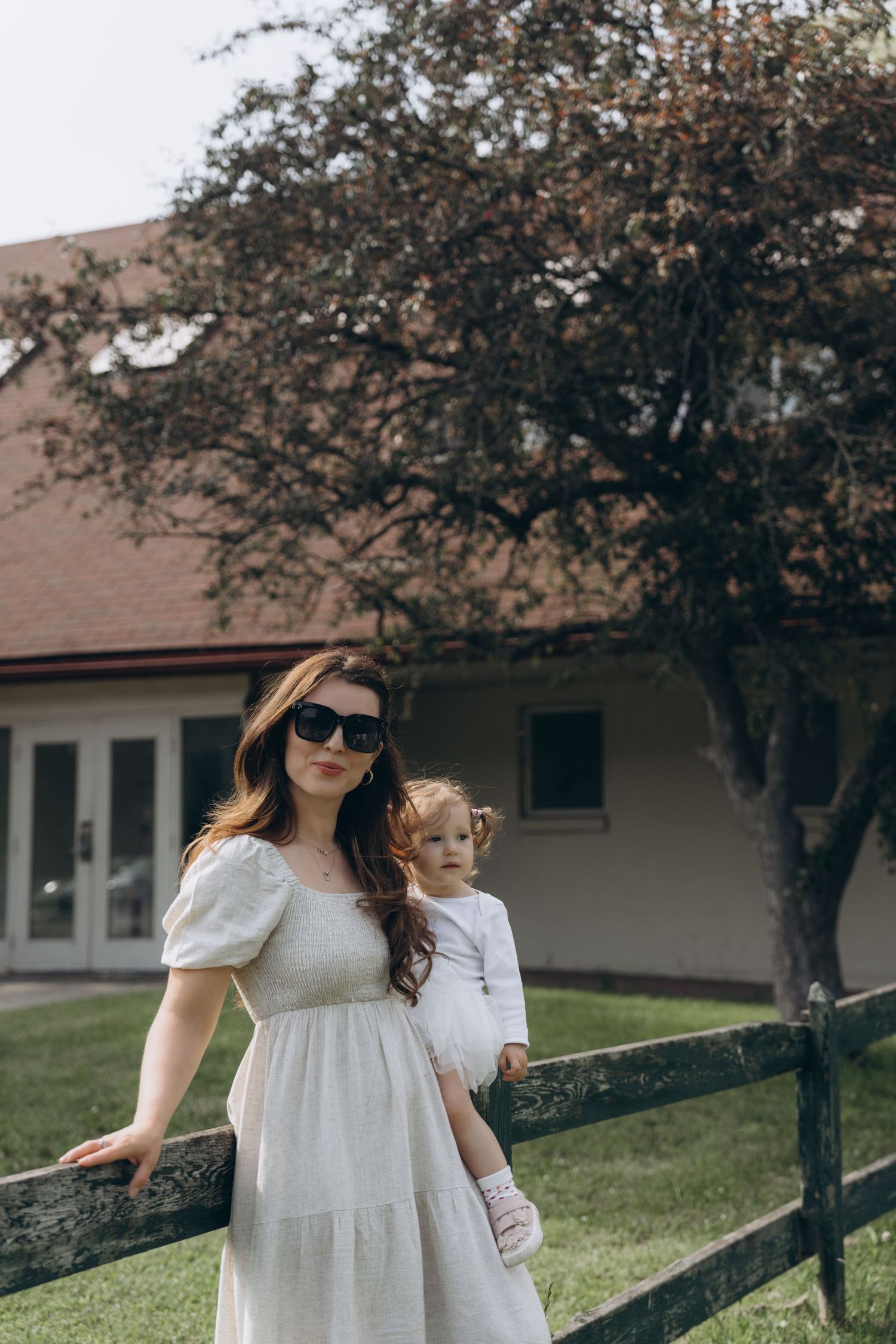Two figures in white dresses stand by a rustic wooden fence near a house with a tree in the background.