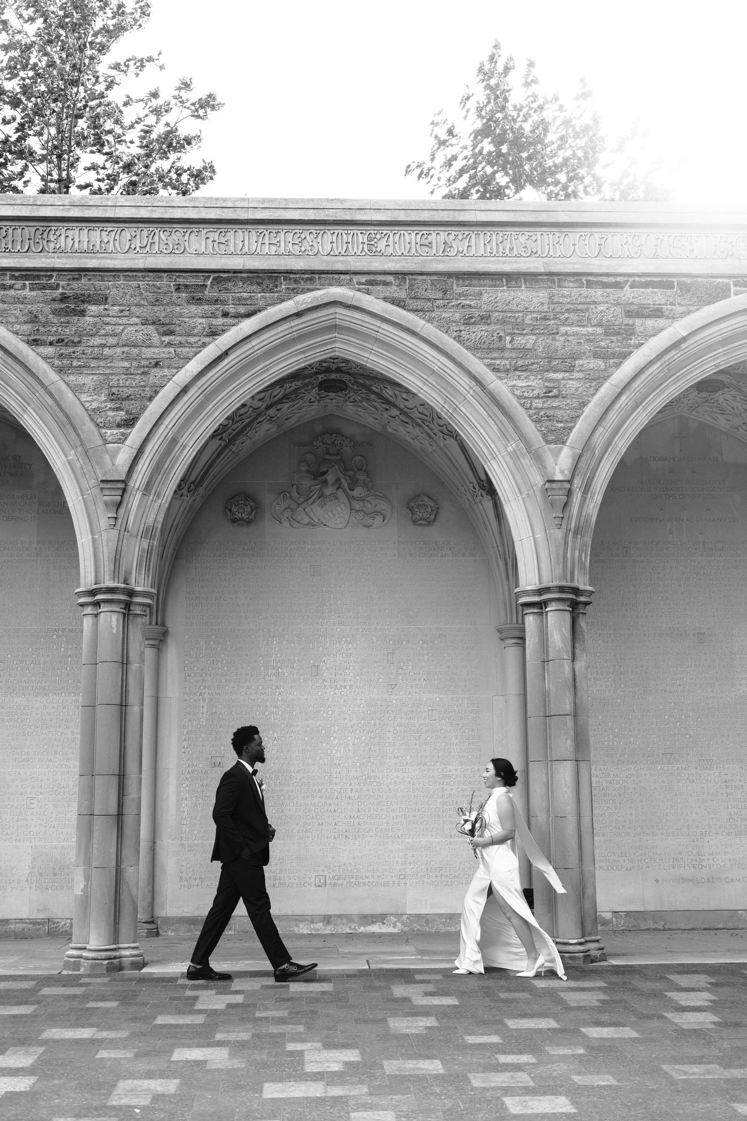 Two people walking in opposite directions under Gothic stone arches in a black and white architectural setting.