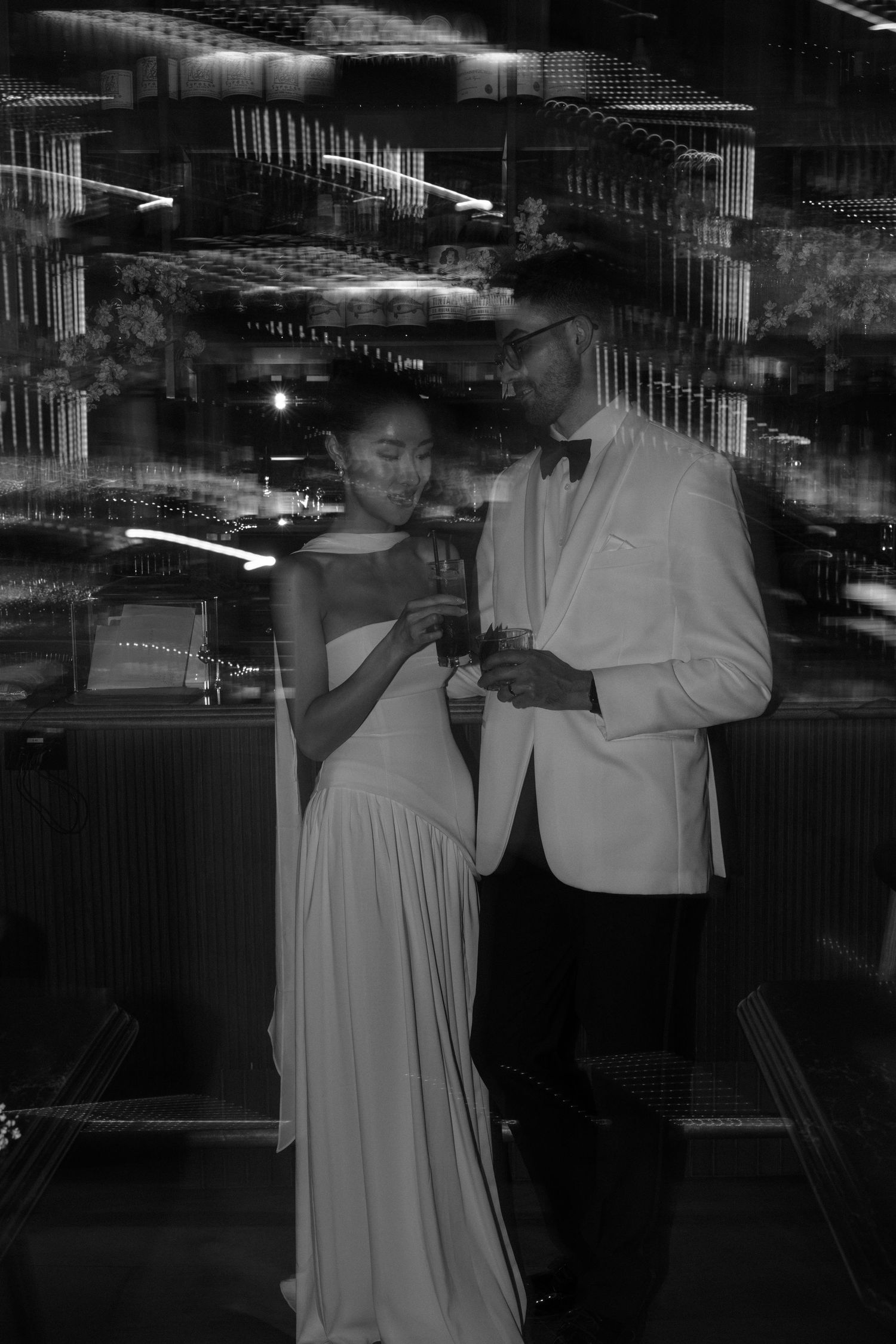 Elegant couple in formal evening attire share a close moment at an upscale venue with reflective surfaces in black and white.