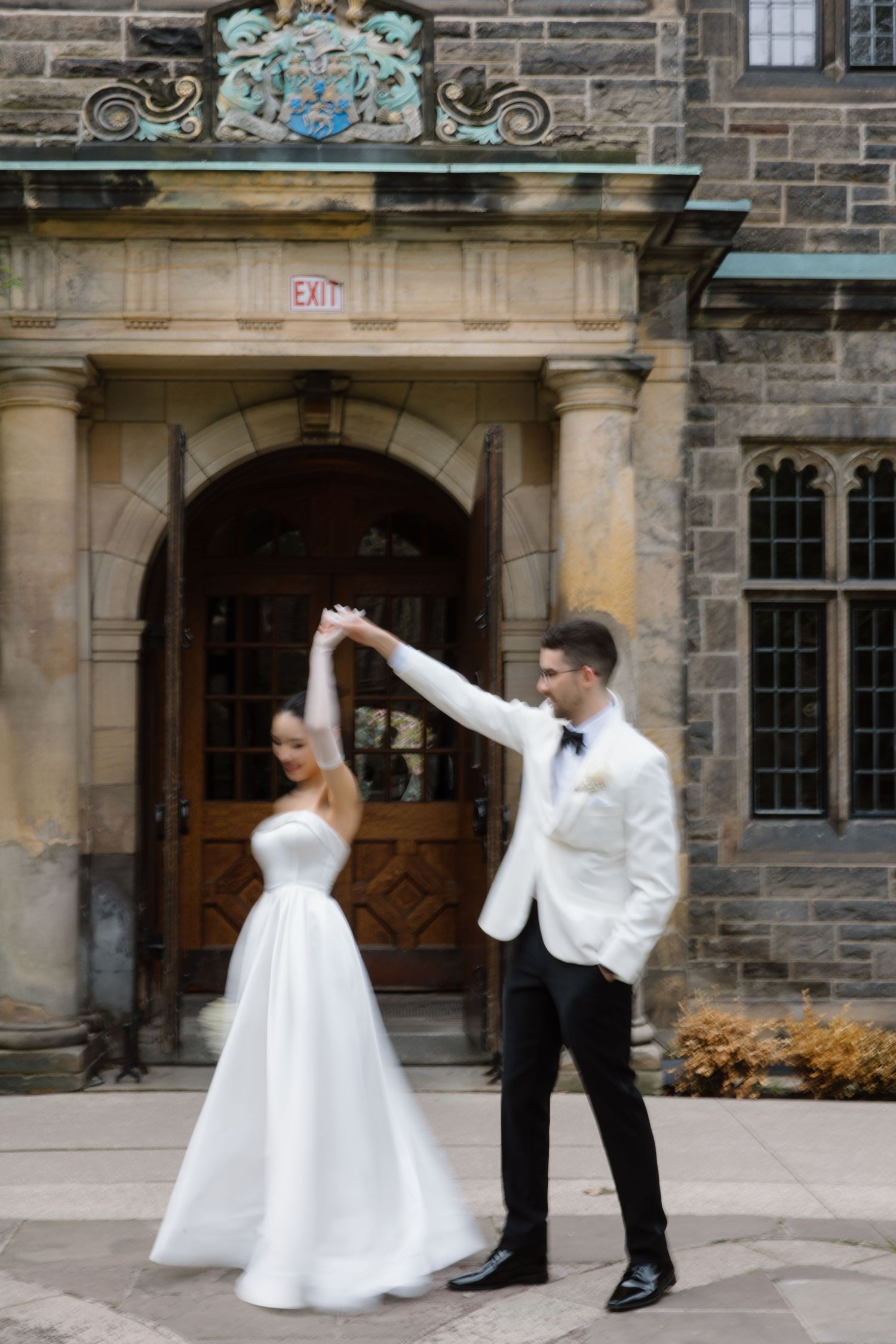An elegant couple in wedding attire dancing and twirling outside a historic stone building with ornate architecture.