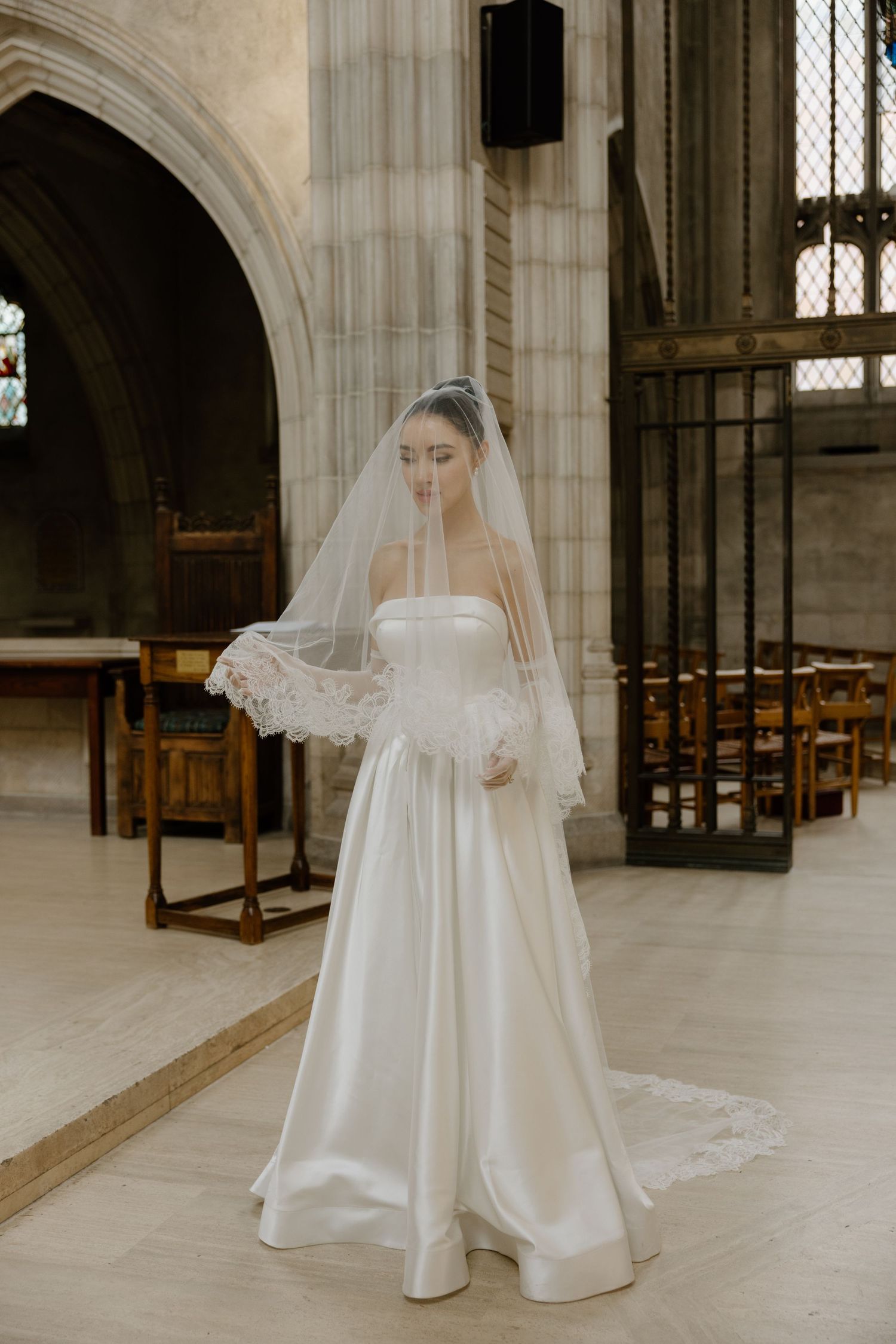A bride in a classic white strapless ball gown and cathedral veil stands in a historic church with stone archways.