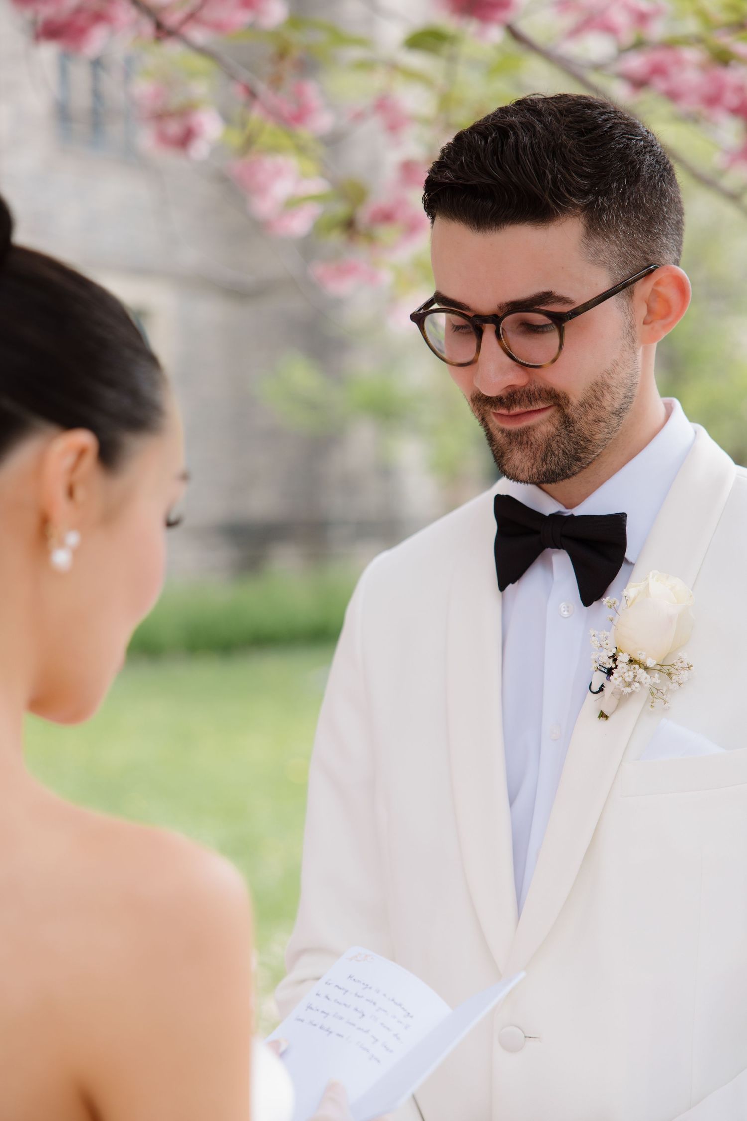 A couple exchanges wedding vows under pink cherry blossoms while the groom wearing glasses and a white tuxedo smiles.