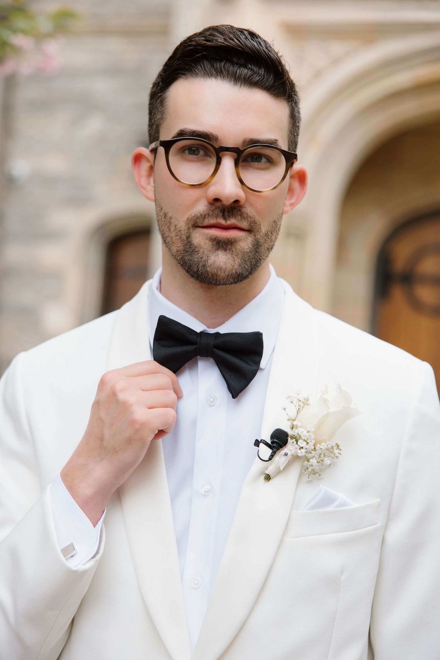 Elegant groom in white tuxedo jacket with black bow tie and glasses adjusts his bowtie while wearing a white boutonniere.