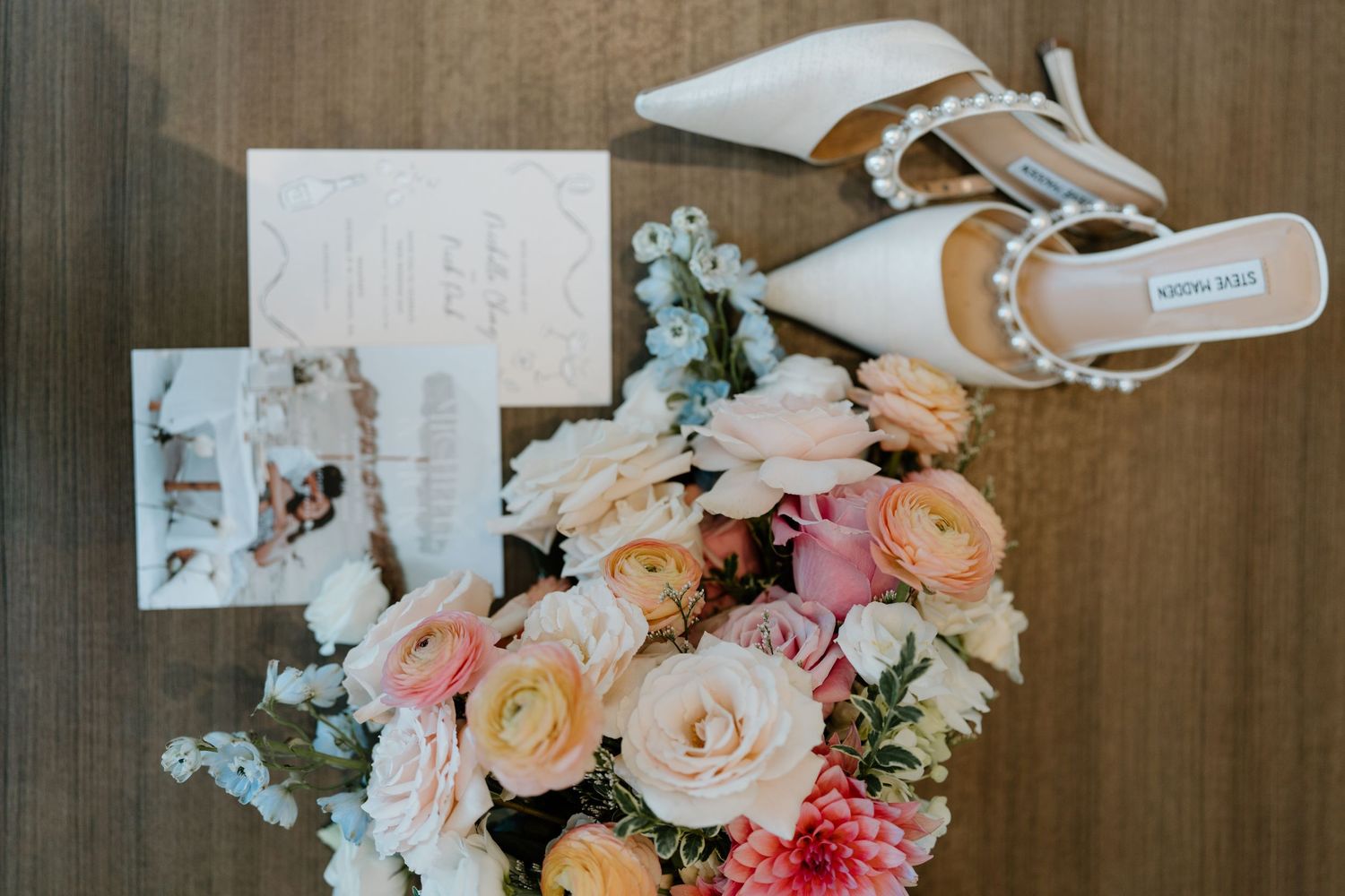Wedding details flat lay featuring bridal shoes, pastel roses and eucalyptus bouquet, and photographs on wooden surface.