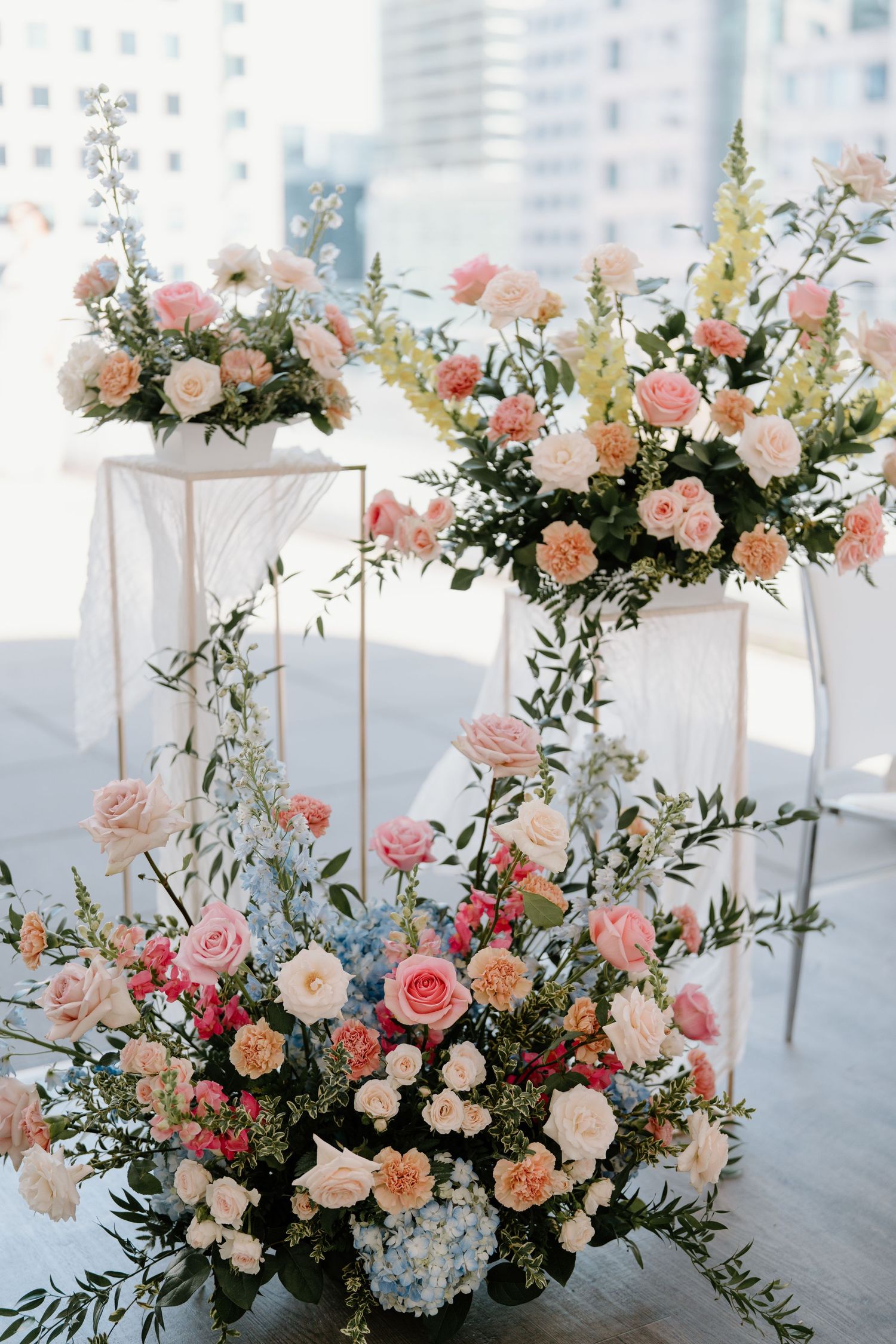 Elegant floral arrangements with pink and peach roses, hydrangeas and greenery displayed on white pedestals at a wedding.