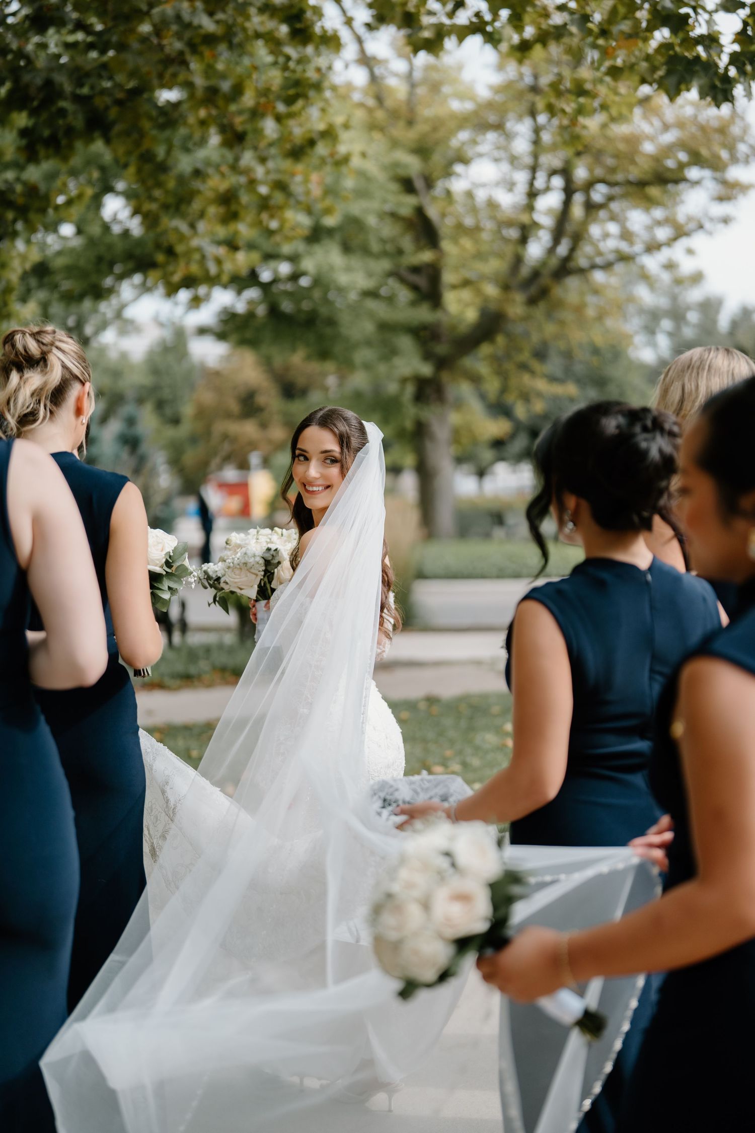 Bridesmaids in navy blue dresses help adjust the bride's flowing white veil during an outdoor wedding photoshoot.