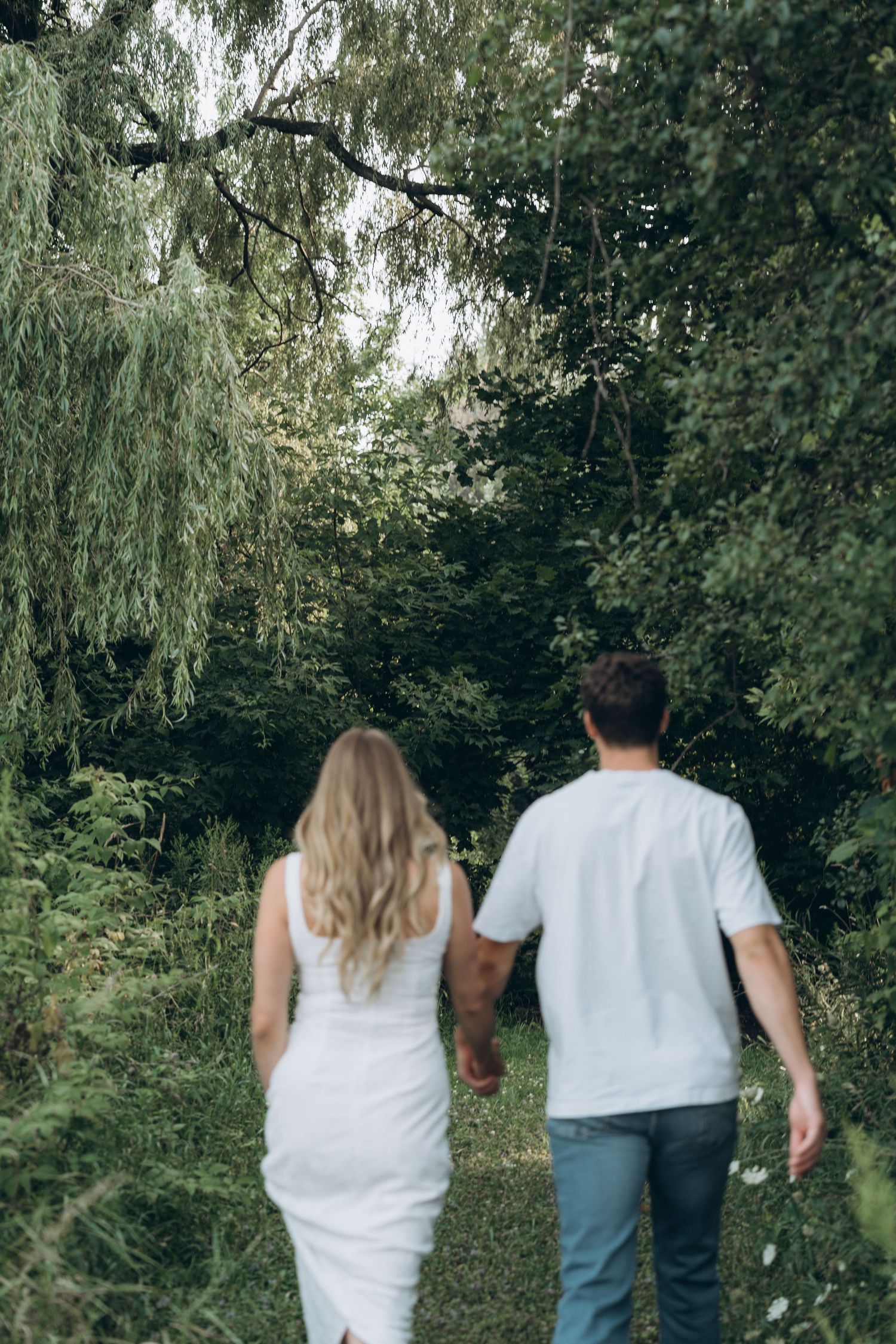 Two people wearing white shirts walk together on a nature path surrounded by green trees and foliage.
