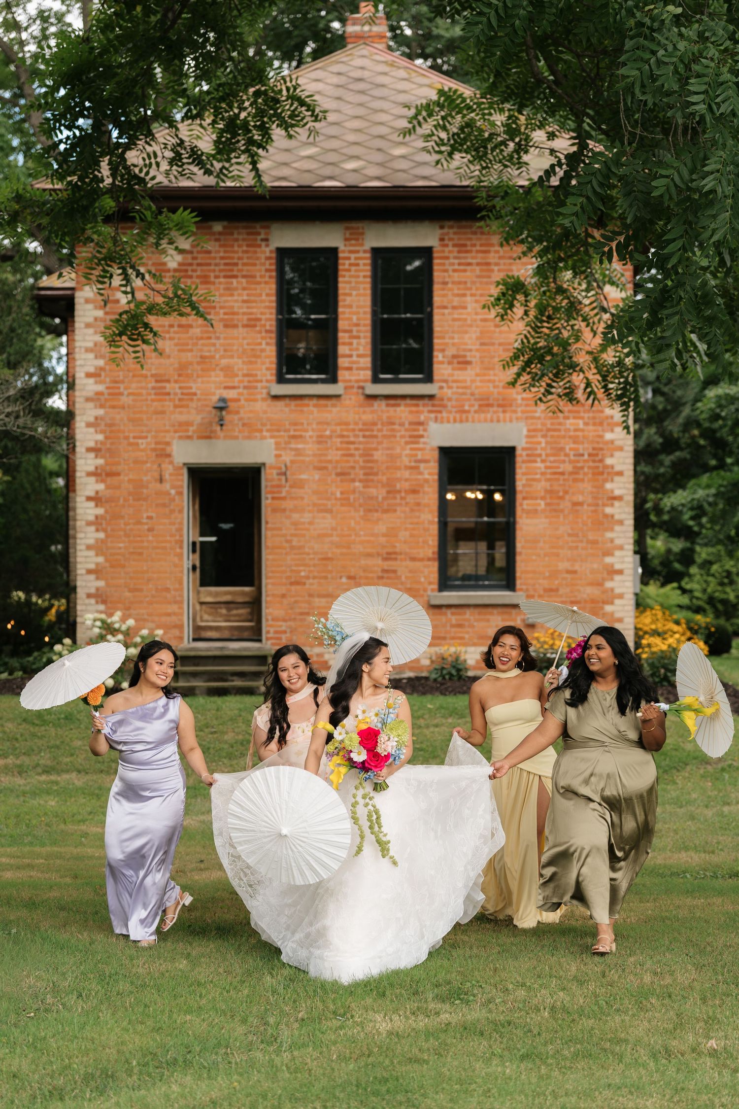 Wedding party poses playfully on grass in front of a rustic brick building with matching lavender dresses.
