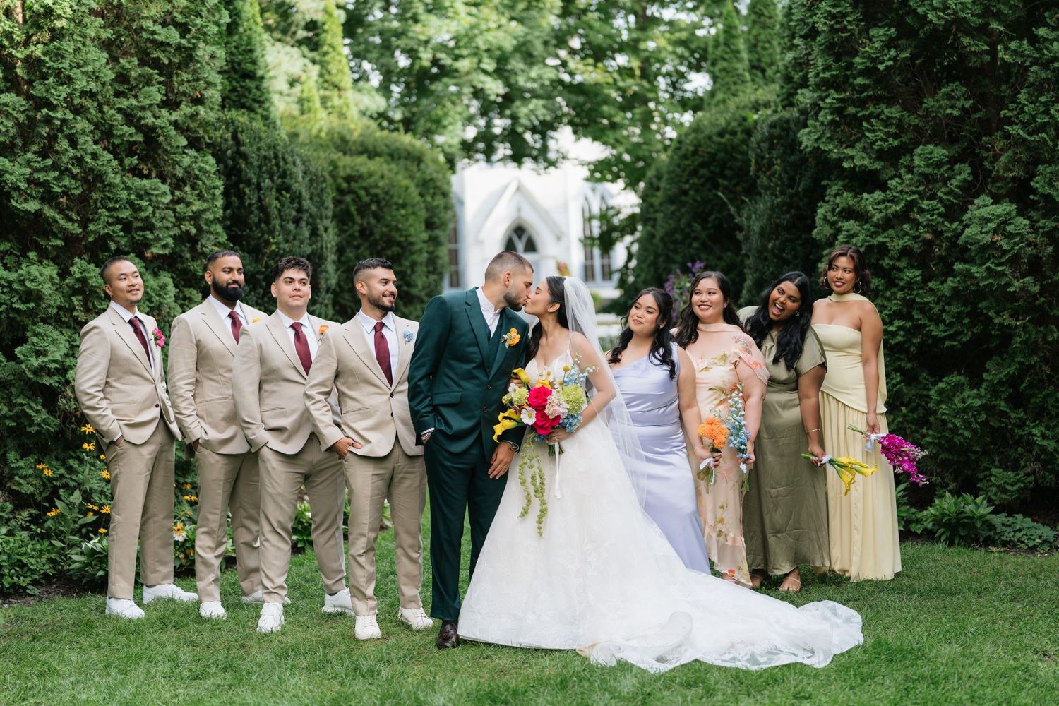 Wedding party in tan suits and champagne dresses poses outdoors in a lush garden setting during summer celebration.