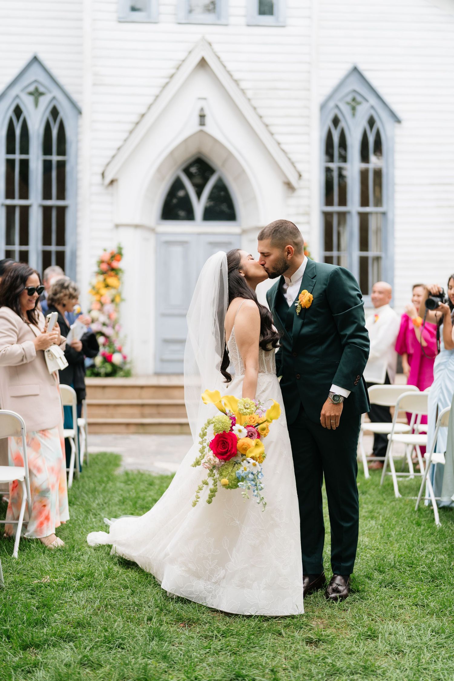 A wedding couple shares an intimate moment on a lawn in front of a white Gothic-style church with arched windows.