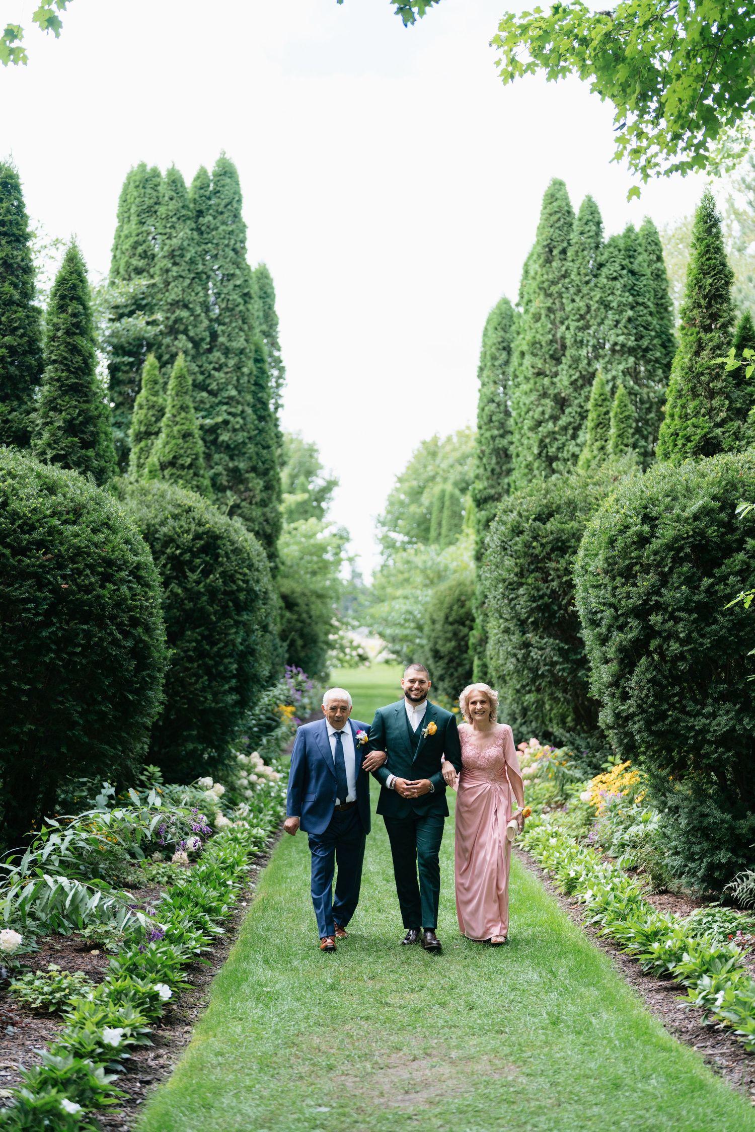 Wedding party walks along a lush garden path lined with tall evergreen trees and colorful flower beds.
