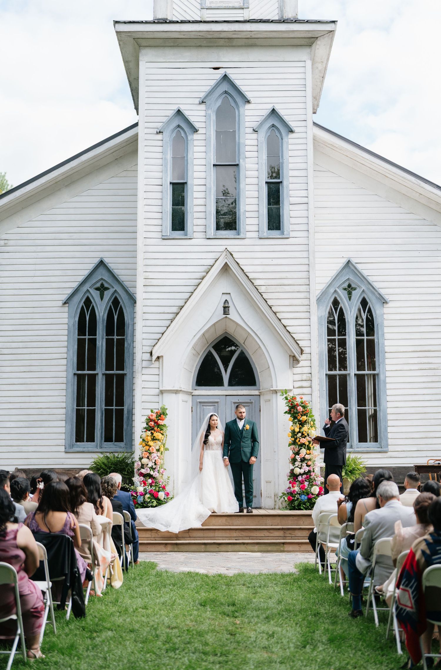 A wedding ceremony takes place outside a charming white Gothic-style church with pointed arch windows and floral decorations.