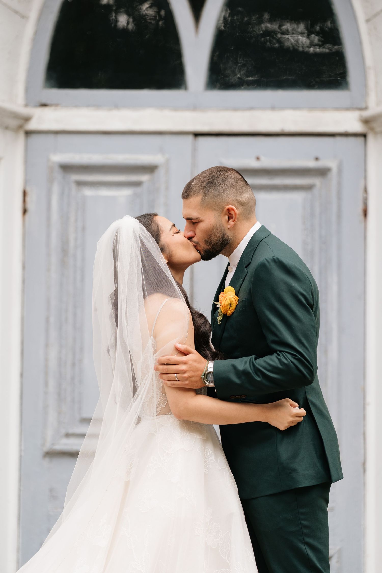 Newlyweds share their first kiss in front of a classic white church door with arched windows.