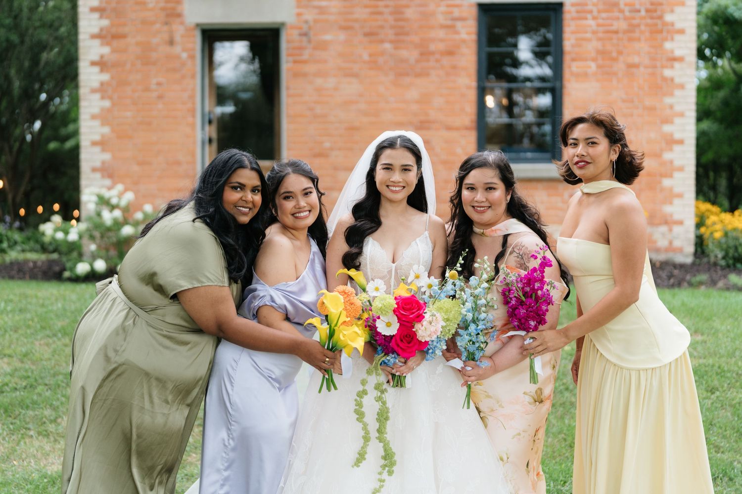 Bridesmaids in pastel dresses standing together outdoors with colorful floral bouquets against brick wall backdrop.