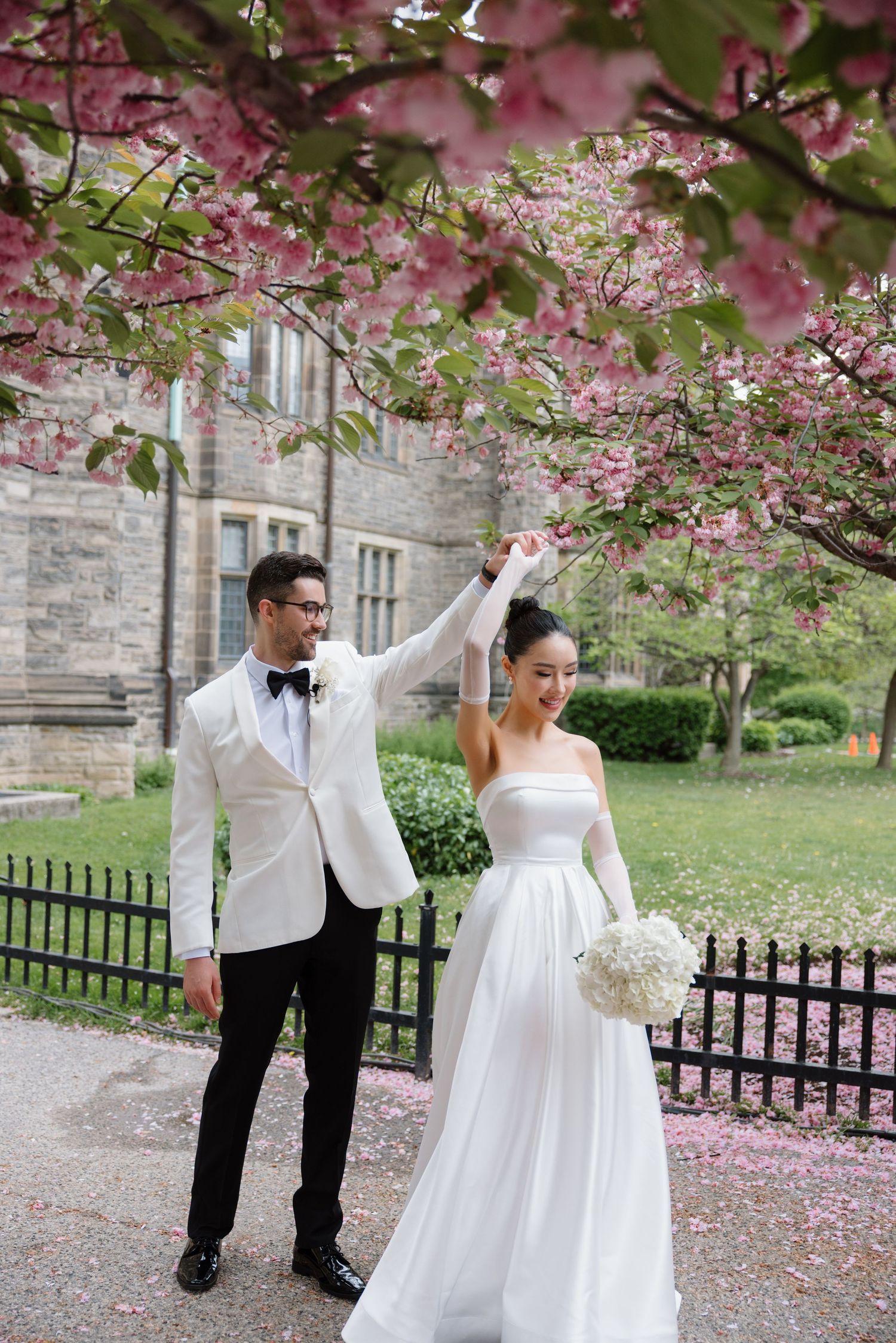 Newlyweds dance under blooming cherry blossom trees near a stone building with pink petals scattered on the ground.