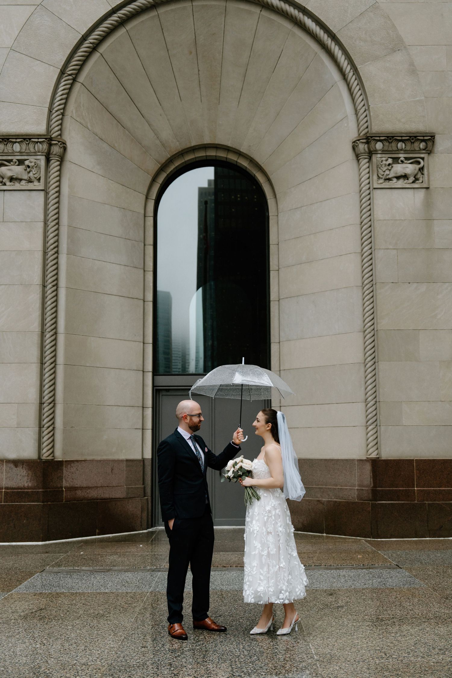 Wedding couple stands under stone arches at historic venue during rainy day celebration.