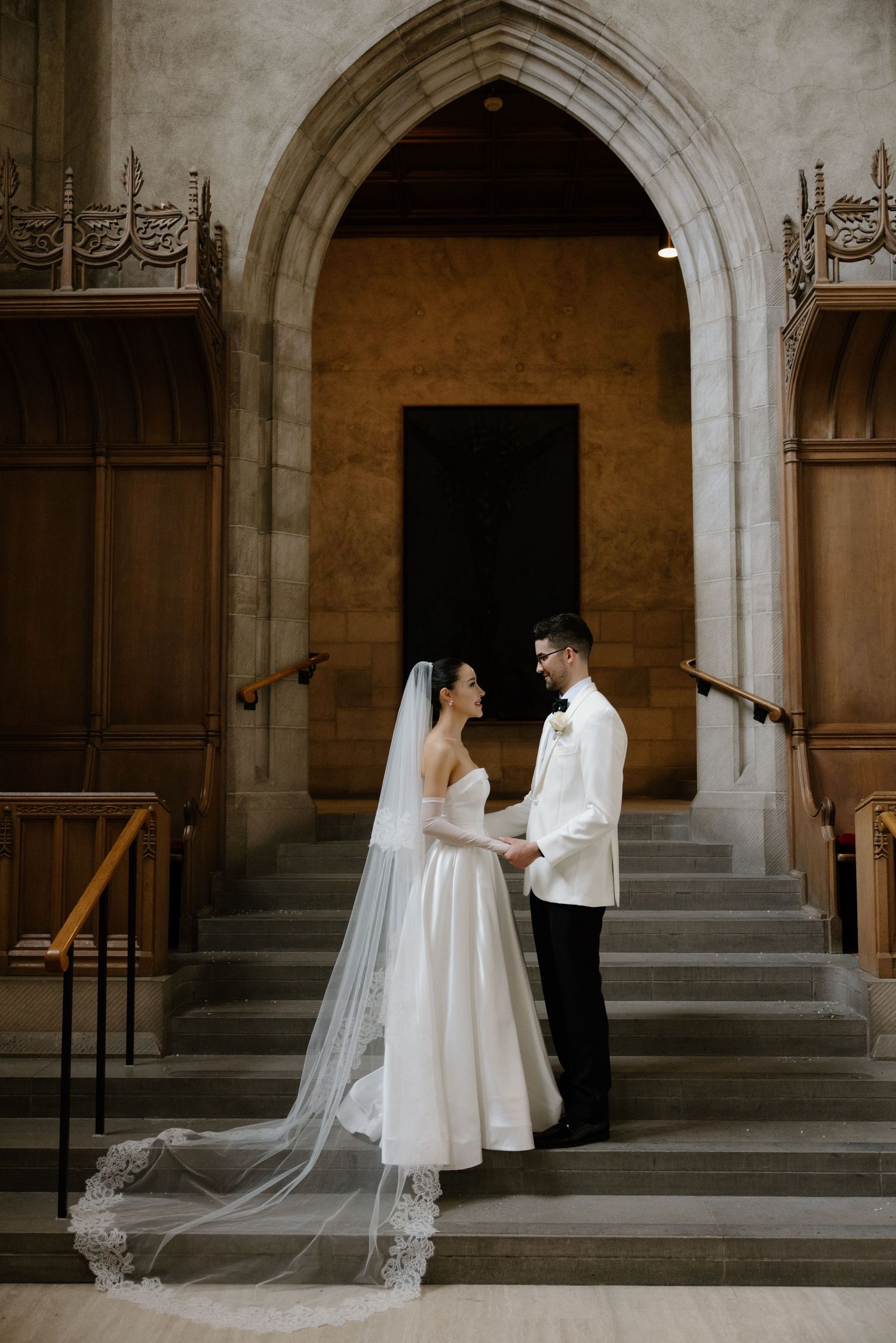 A couple stands together on stone steps beneath a Gothic archway, with the bride wearing a flowing lace veil and white gown.