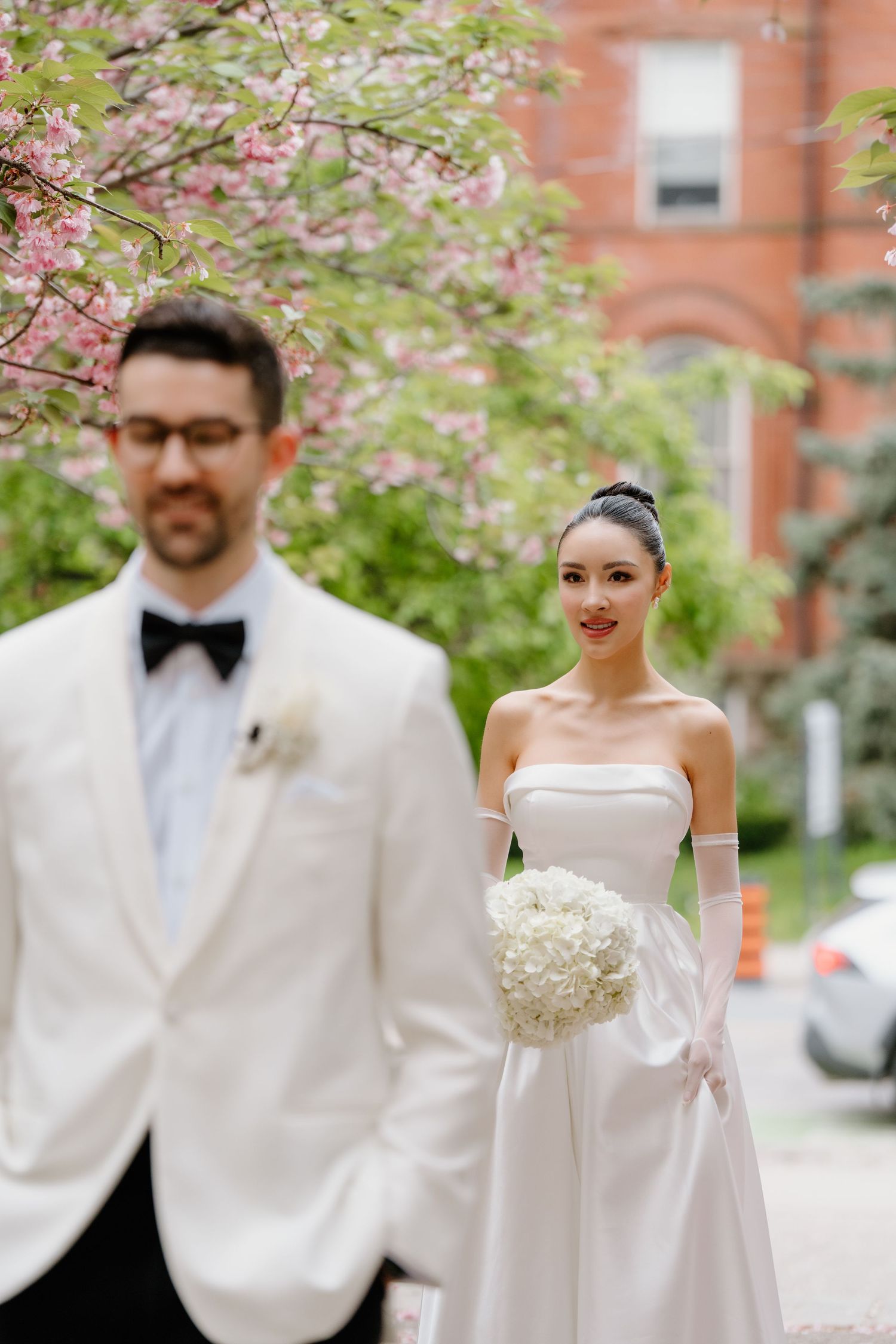 A couple in wedding attire stands beneath pink cherry blossoms with a classic brick building in the background.