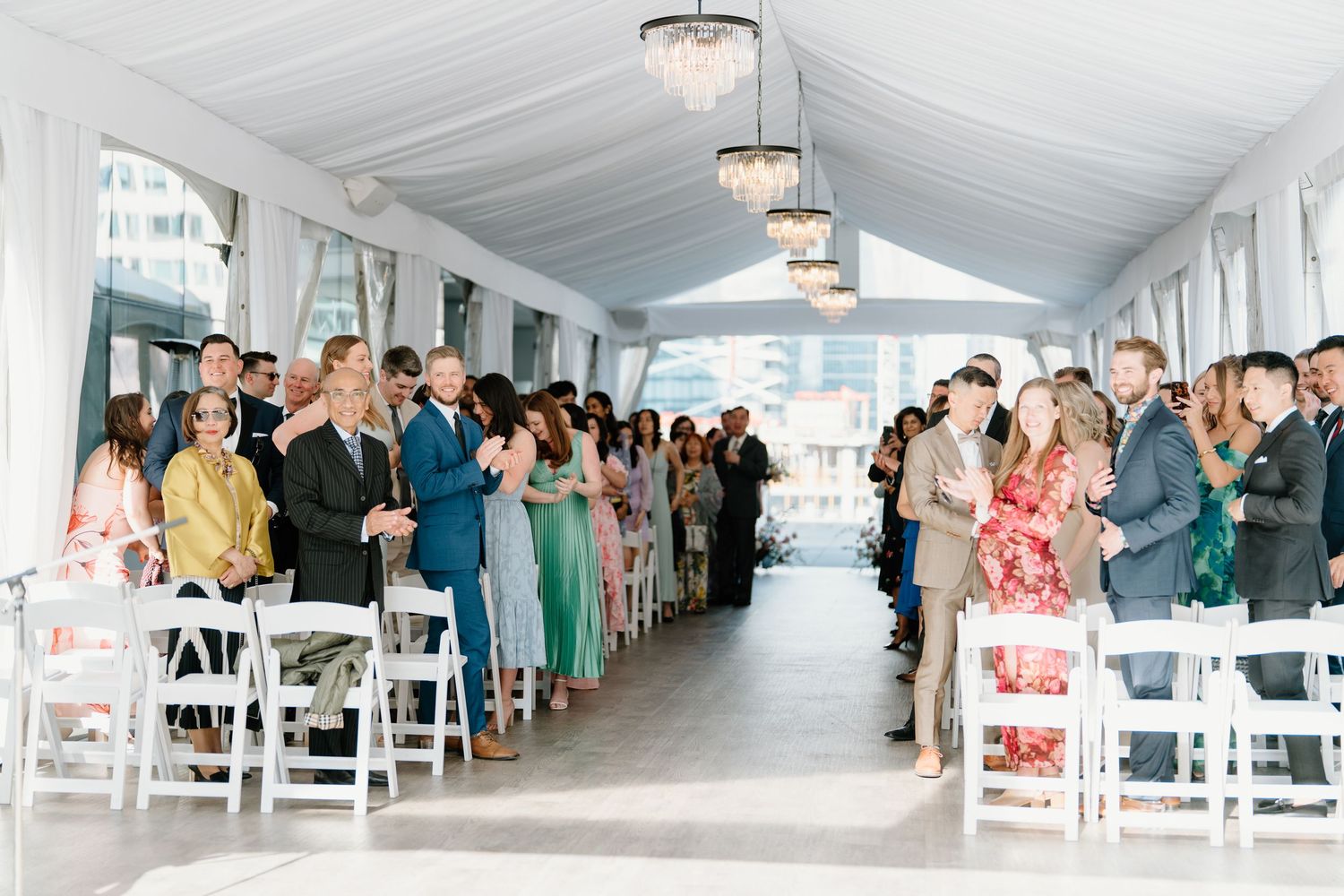 Guests stand in a white decorated wedding tent during a ceremony with chandeliers overhead.
