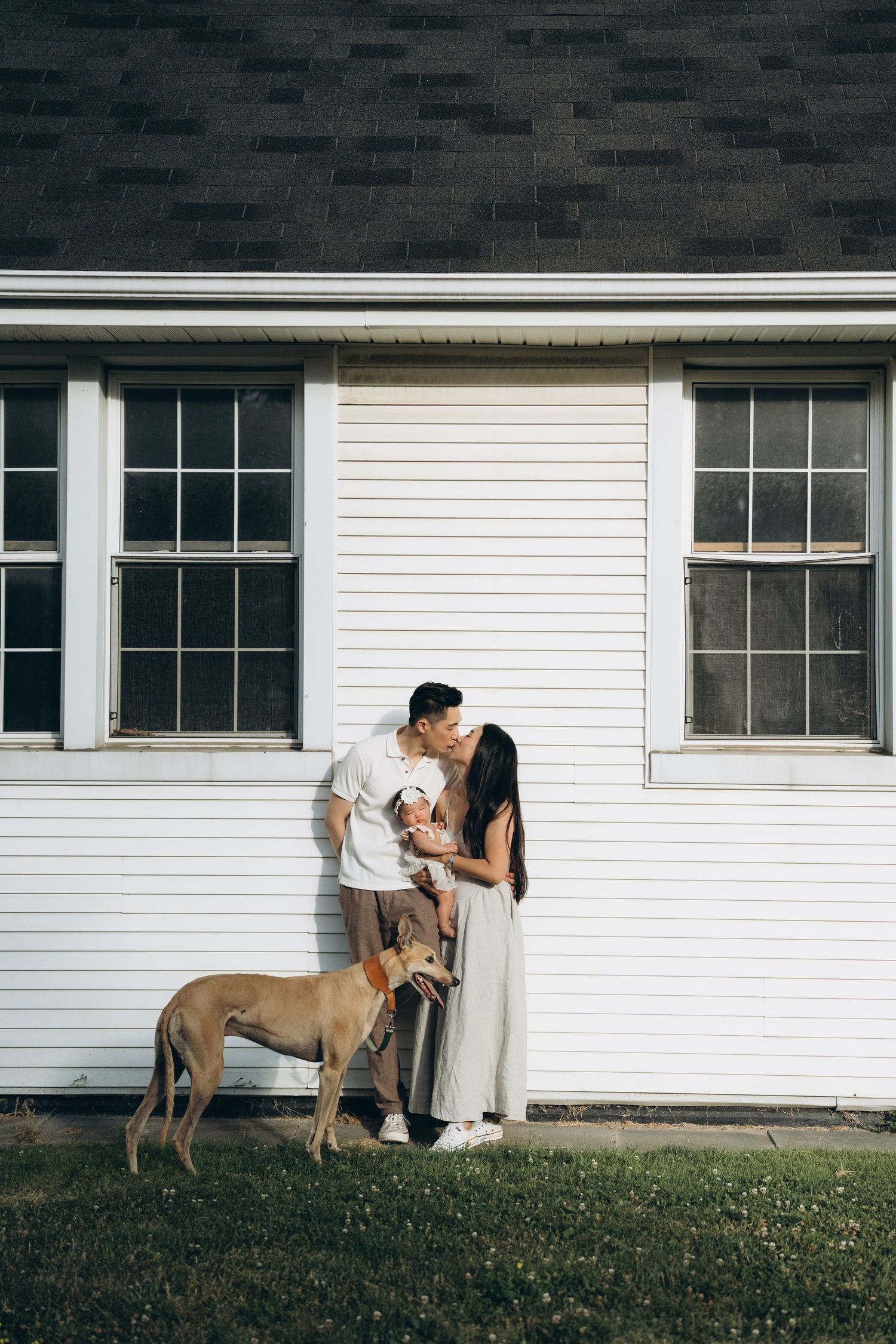 A couple embraces by a white clapboard house while their dog stands beside them.