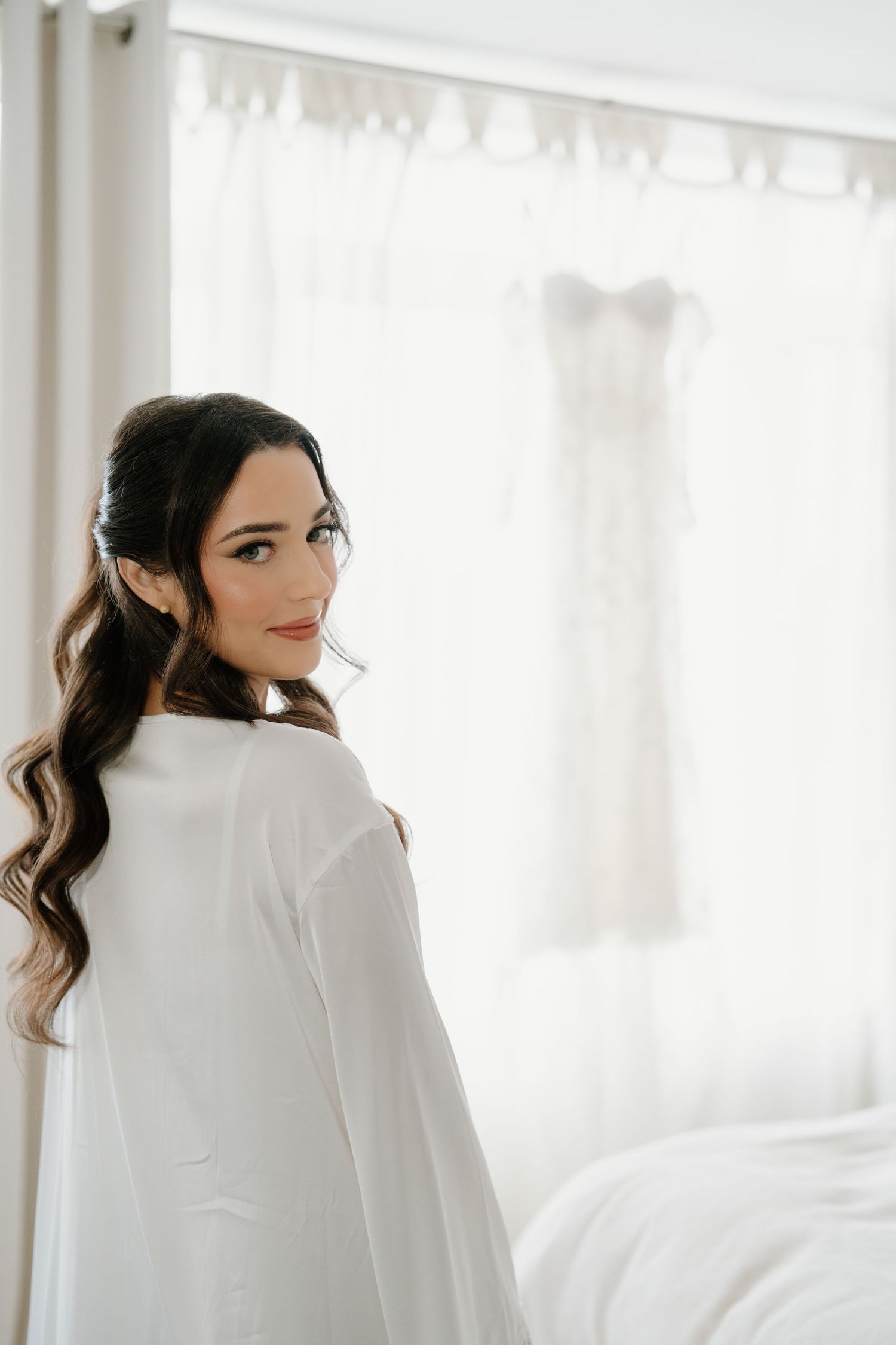 A figure wrapped in a white blanket sits on a bed in a bright, sunlit room with sheer curtains.